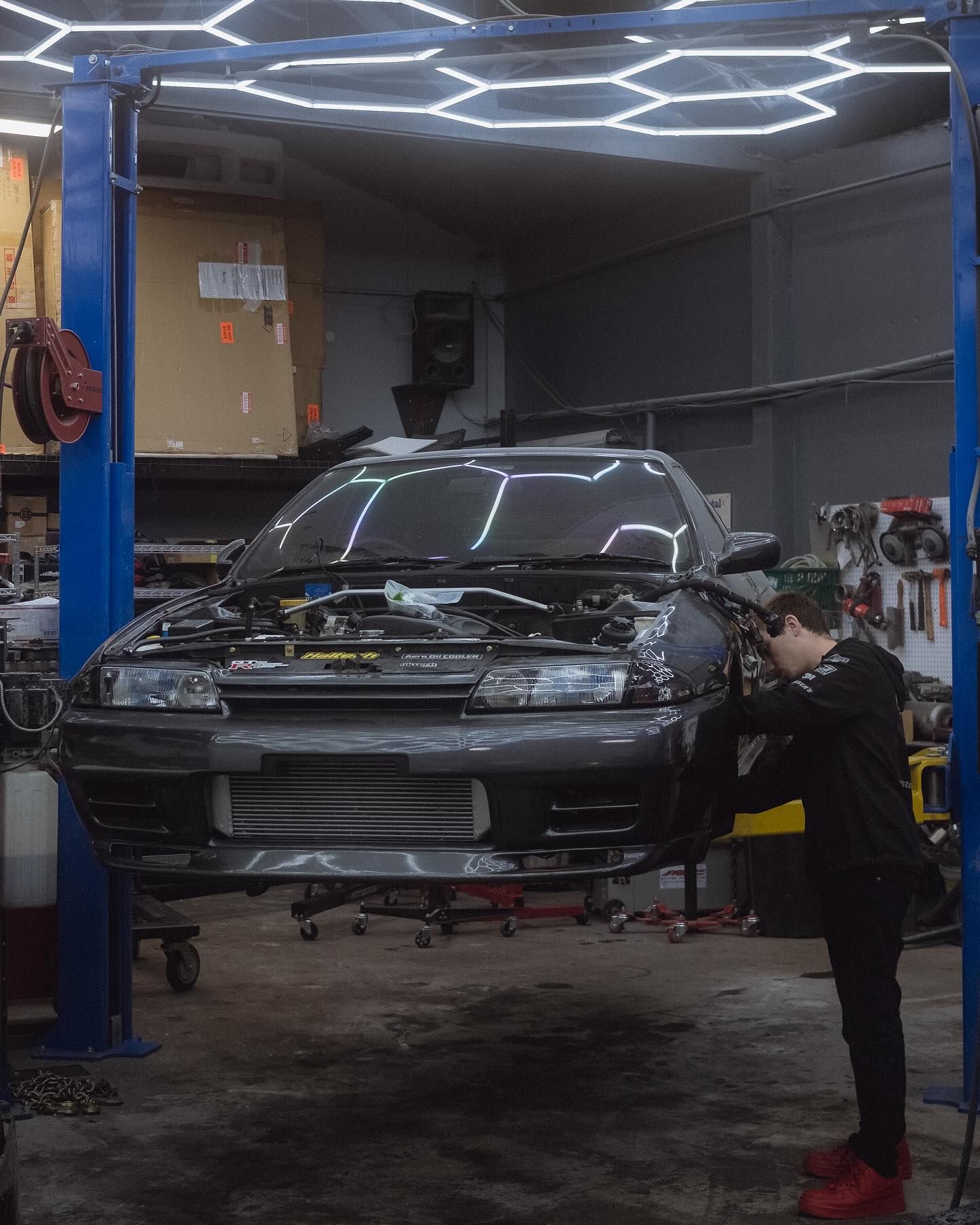 Car being worked on in a garage by a person, car on a lift, shop tools visible.