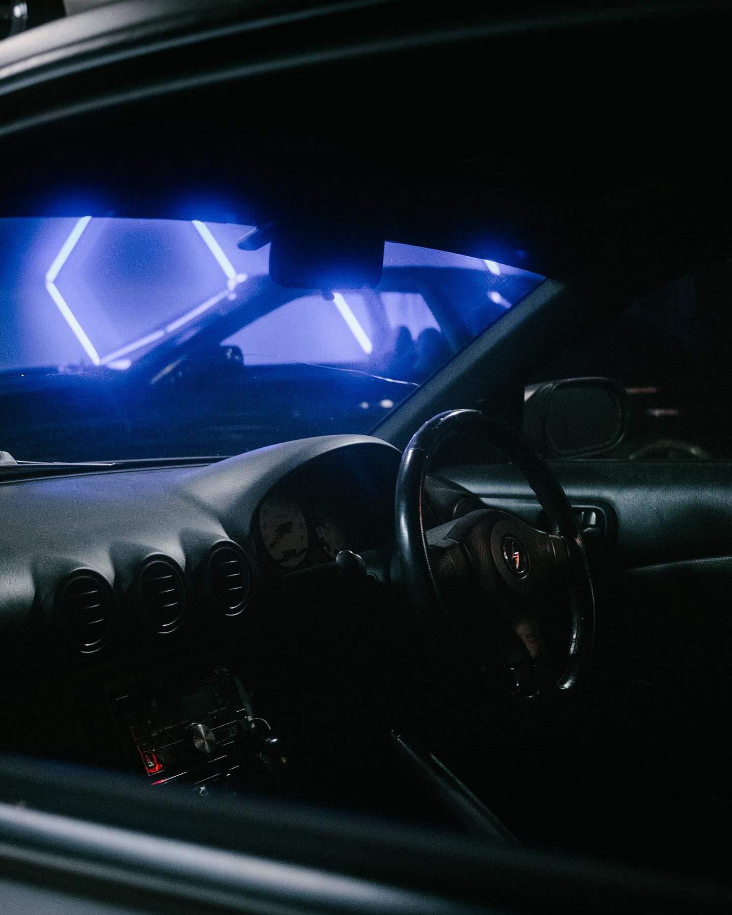 Dark interior of a car, view through the window. Steering wheel visible, neon blue lights outside.