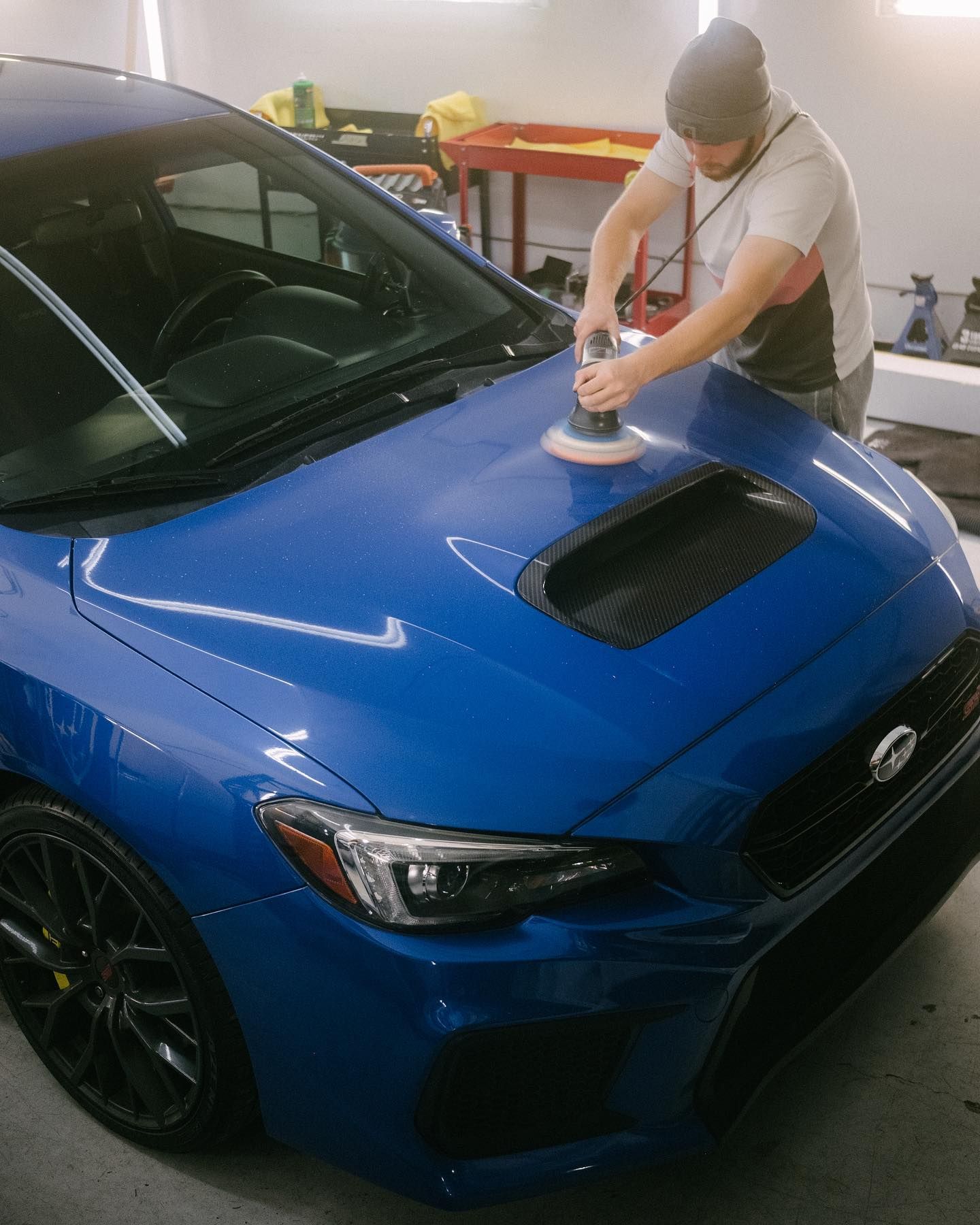 A person polishing the hood of a blue Subaru car with a machine in a workshop.