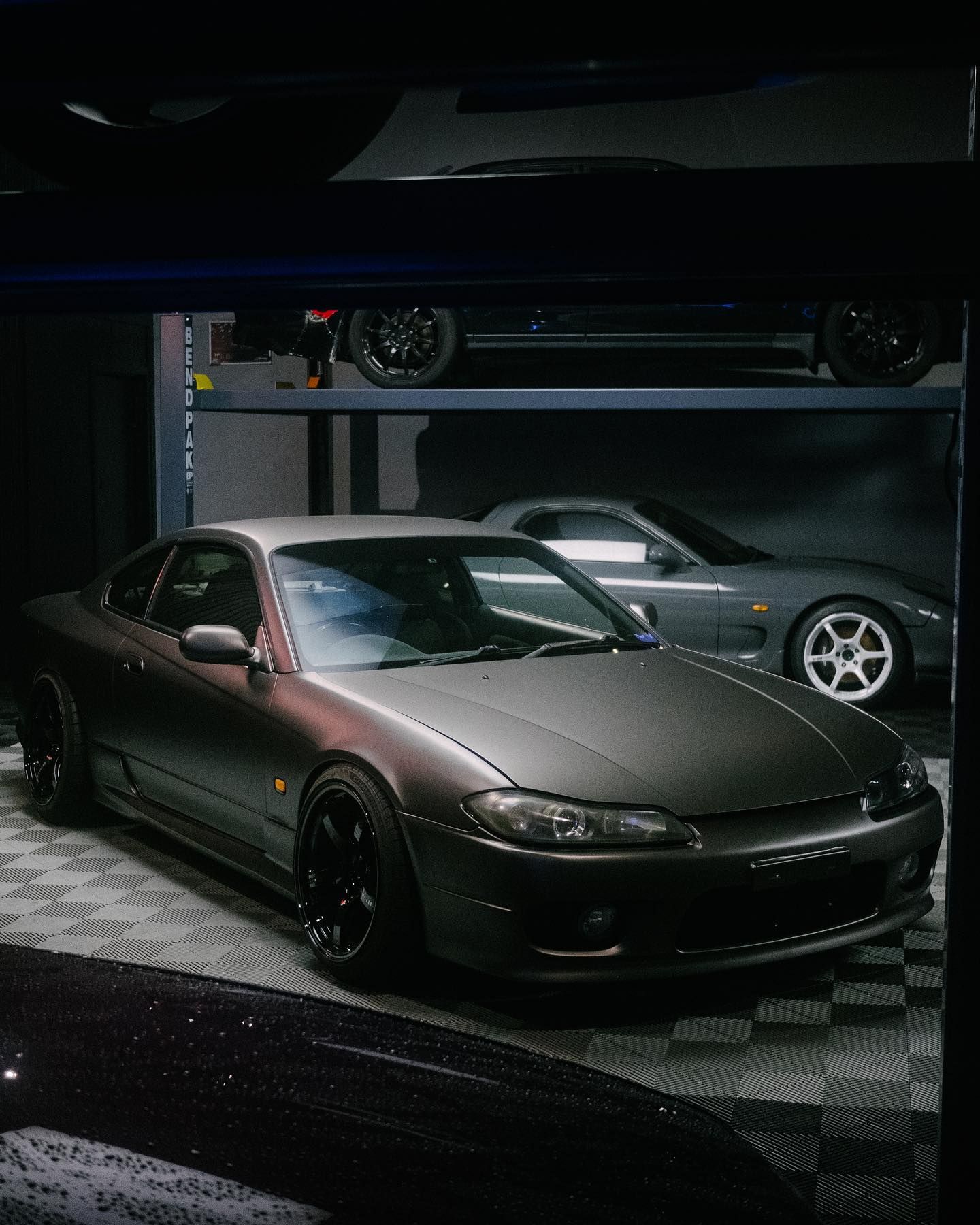 Dark gray sports car in a garage with another on a lift; dim lighting.