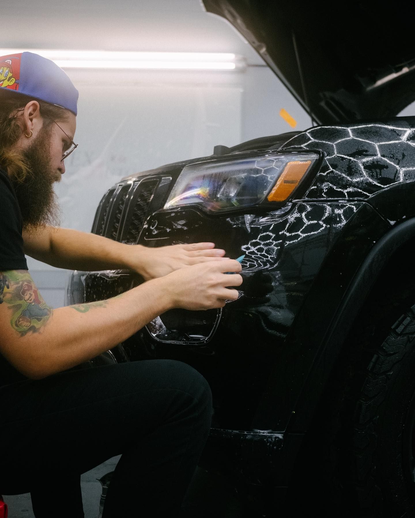 Person applying protective film to a black car's front bumper, inside a workshop.