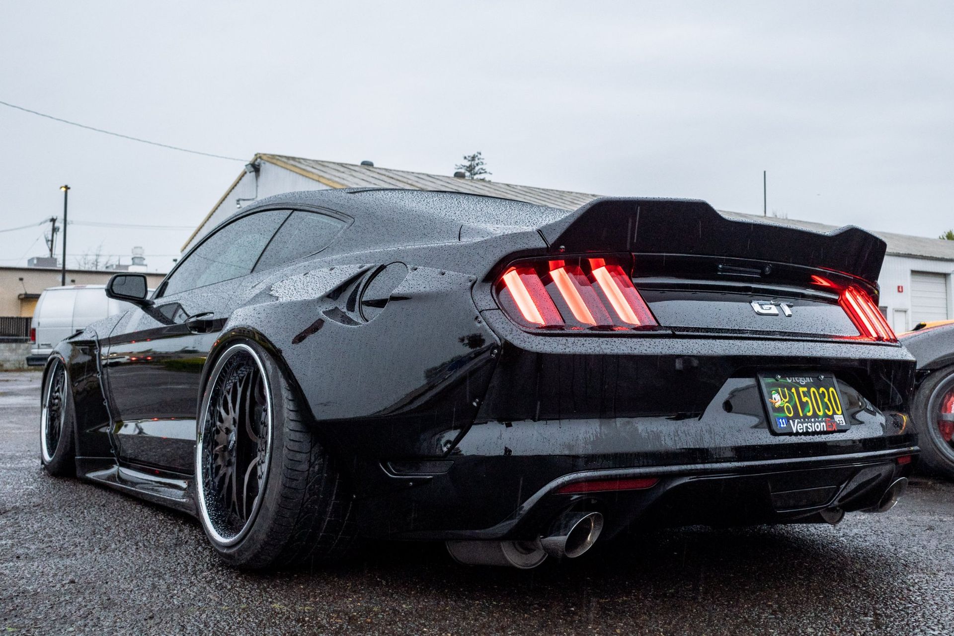 Black Mustang sports car with aftermarket wide body kit, rear view, wet asphalt.