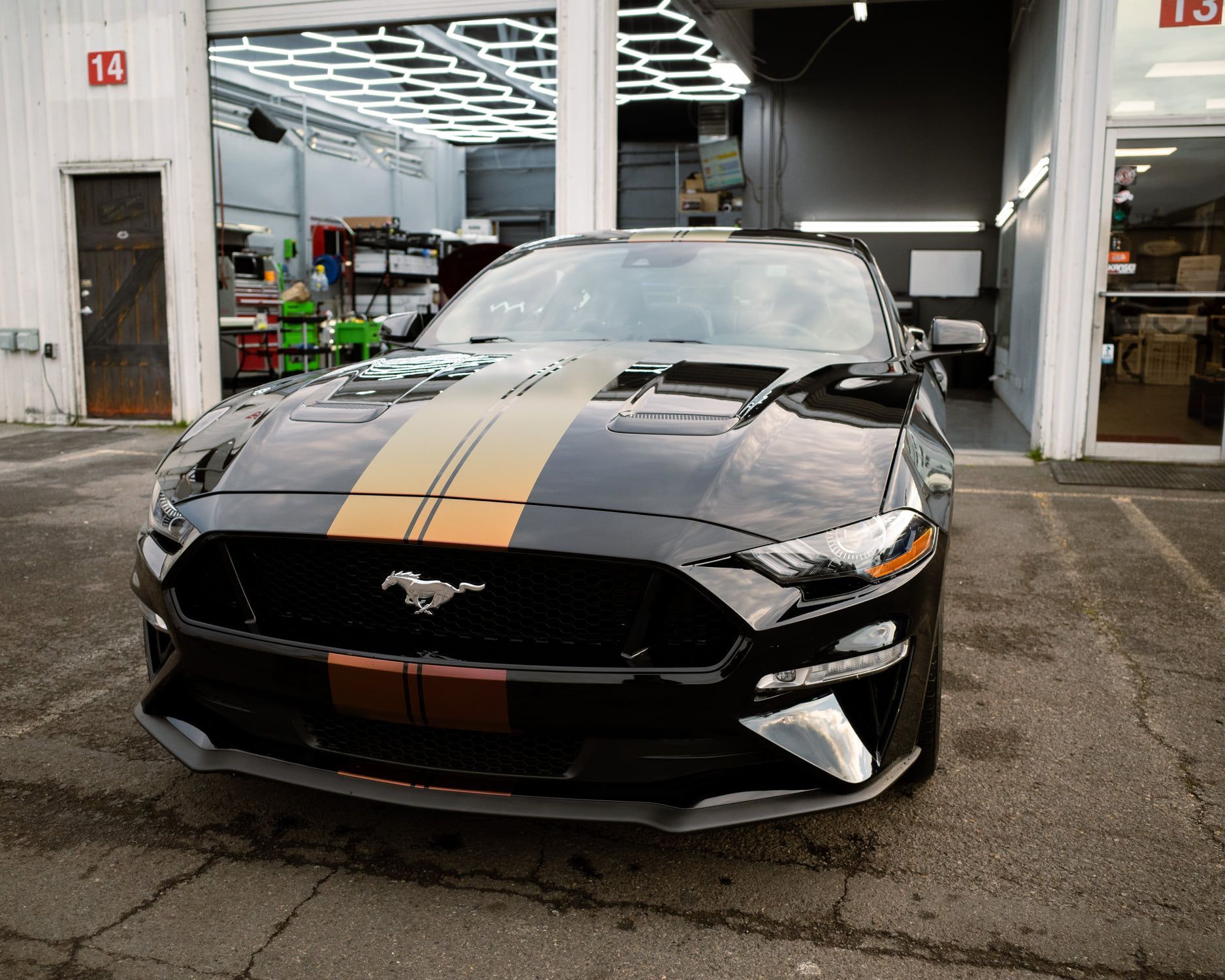 Black Ford Mustang with gold racing stripes parked in front of a garage.