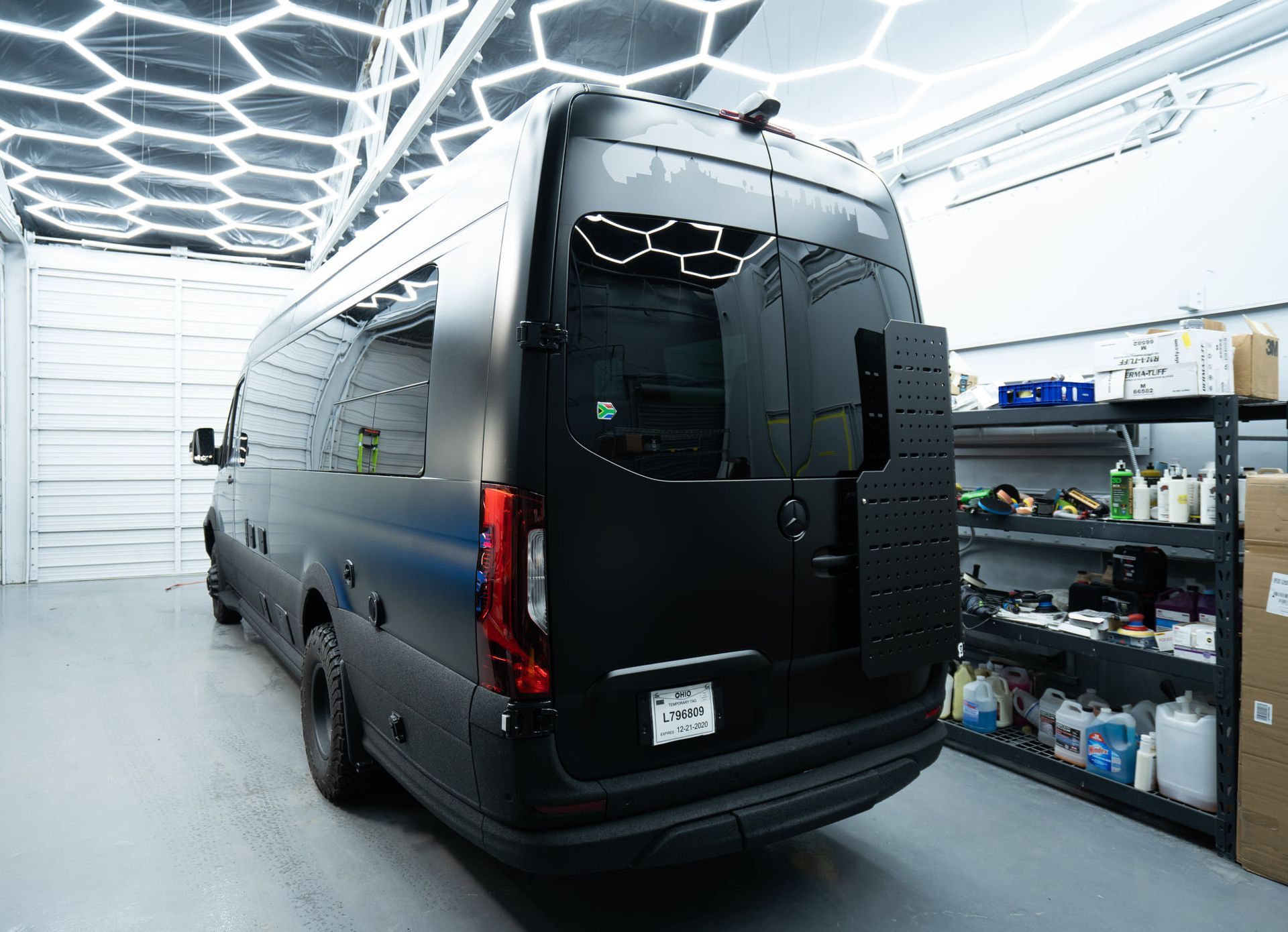 Black van with rack on back parked inside a well-lit garage, various items on shelves to the right.
