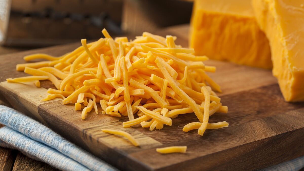 Shredded cheddar cheese on a wooden board with a block of cheese and a grater.