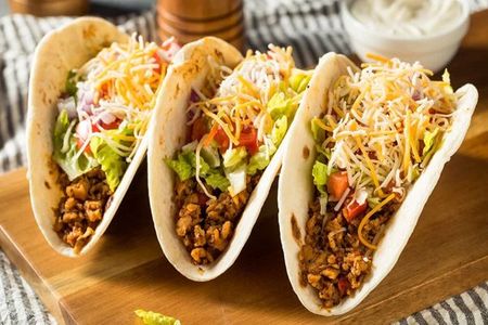 Three fully-loaded tacos on a wooden board, with ground meat, lettuce, cheese, and tomatoes.