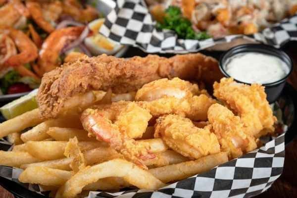 Fried fish, shrimp, and french fries in a basket with dipping sauce; other seafood visible in the background.
