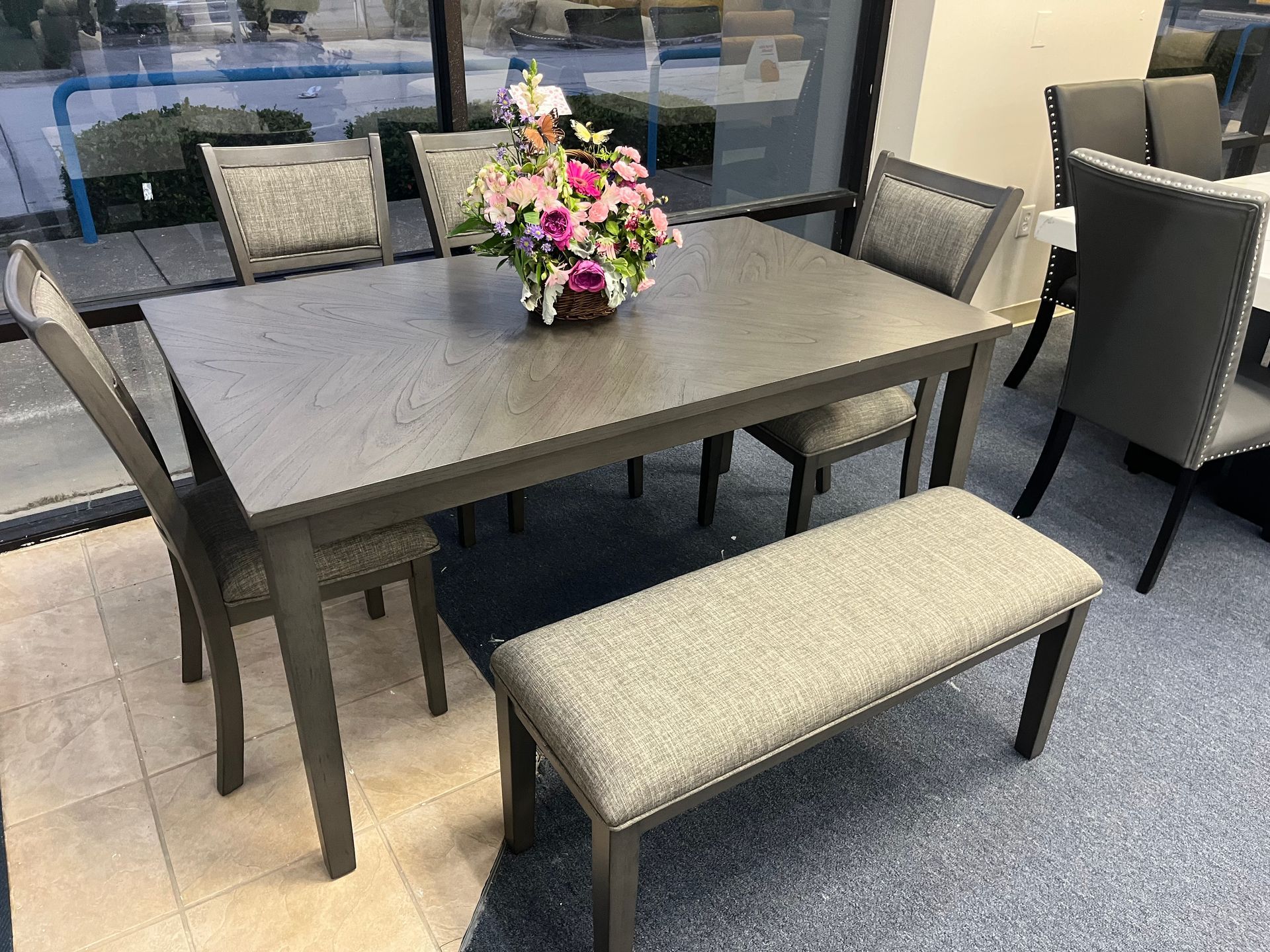 Patio dining set with gray table, four chairs, matching bench, and flower centerpiece near a window.