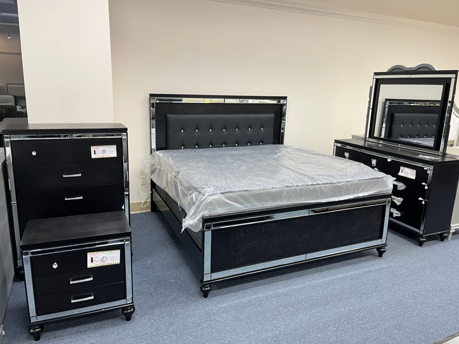 Bedroom furniture set with black bed, dresser, nightstand, and mirror on a carpeted showroom floor