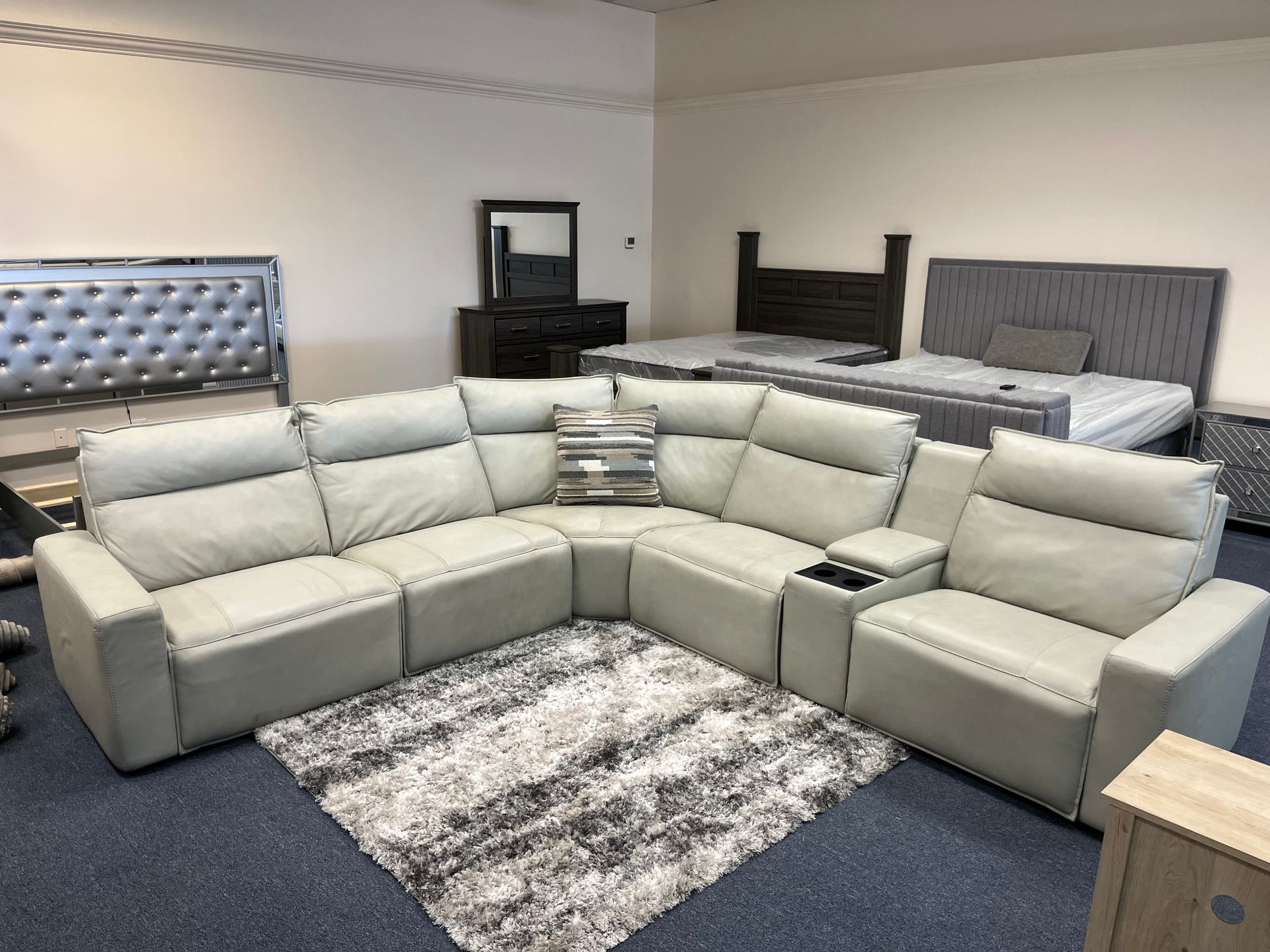 U-shaped beige sectional in a showroom with beds, dresser, and rug in the background