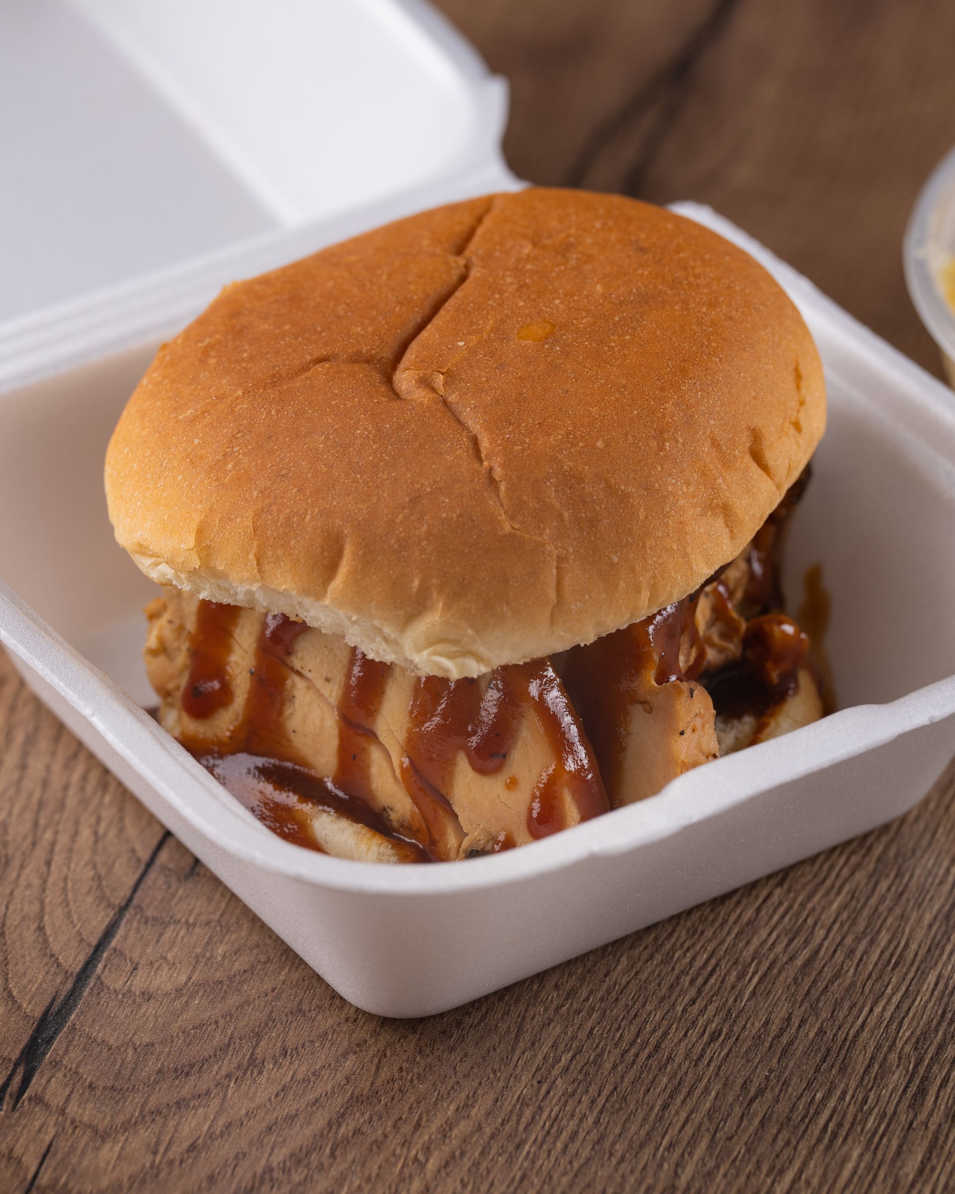 Pulled pork sandwich with barbecue sauce in a white takeout container on wood surface.
