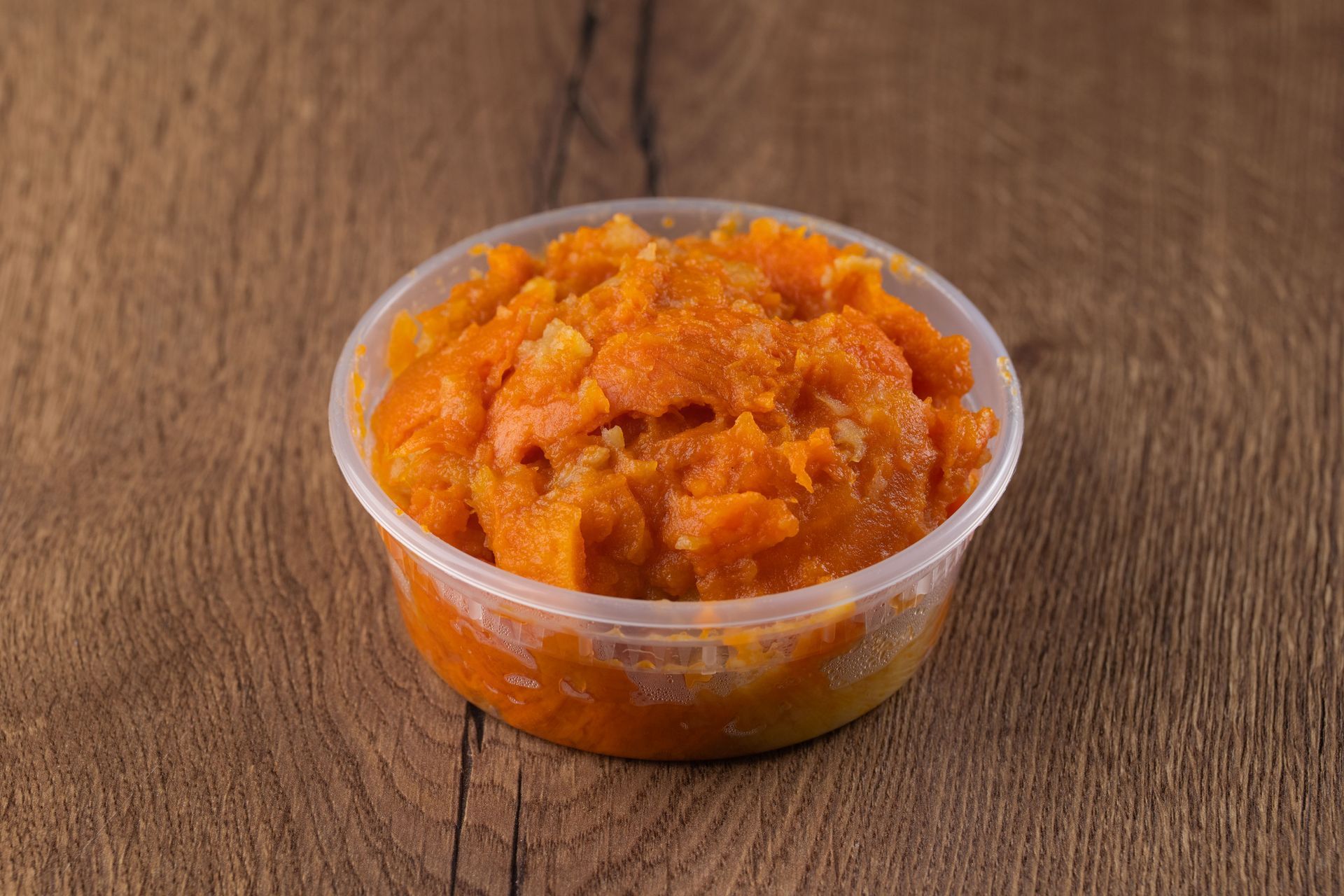Orange food, possibly mashed sweet potatoes, in a clear plastic container on wood.