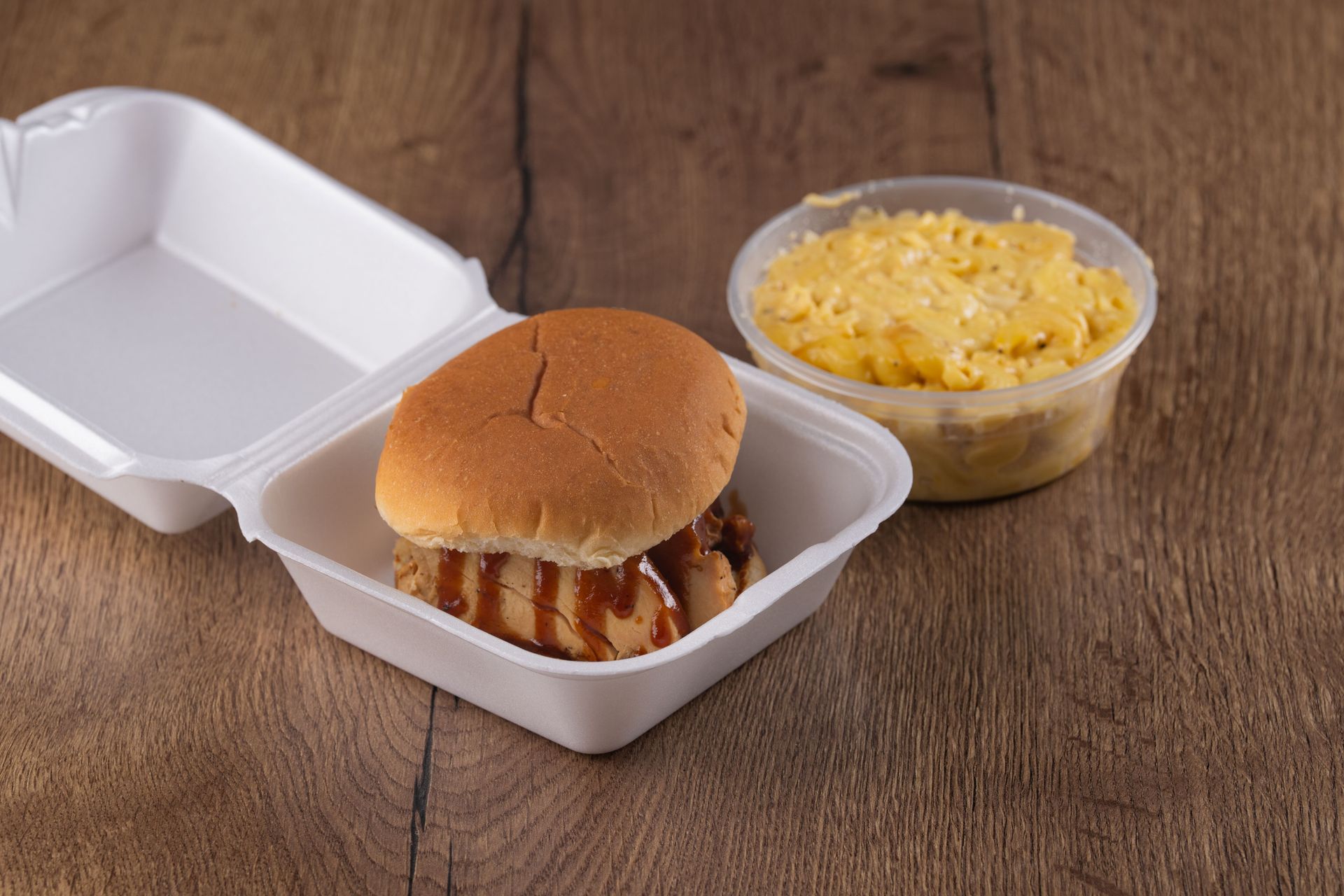 BBQ sandwich with mac and cheese in a styrofoam container, on a wood surface.