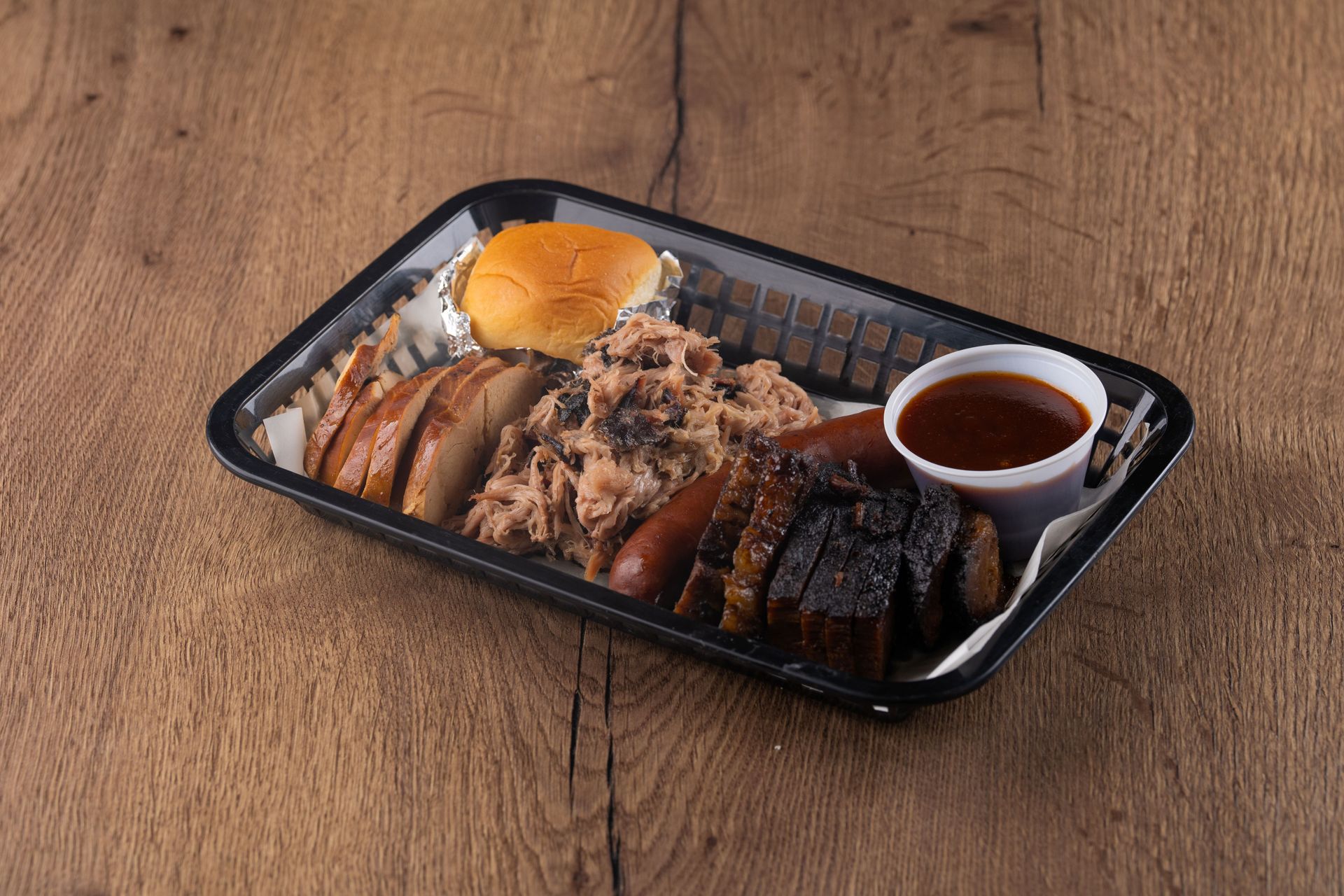 Barbecue platter with brisket, sausage, pulled pork, a bun, and sauce, on a wooden table.