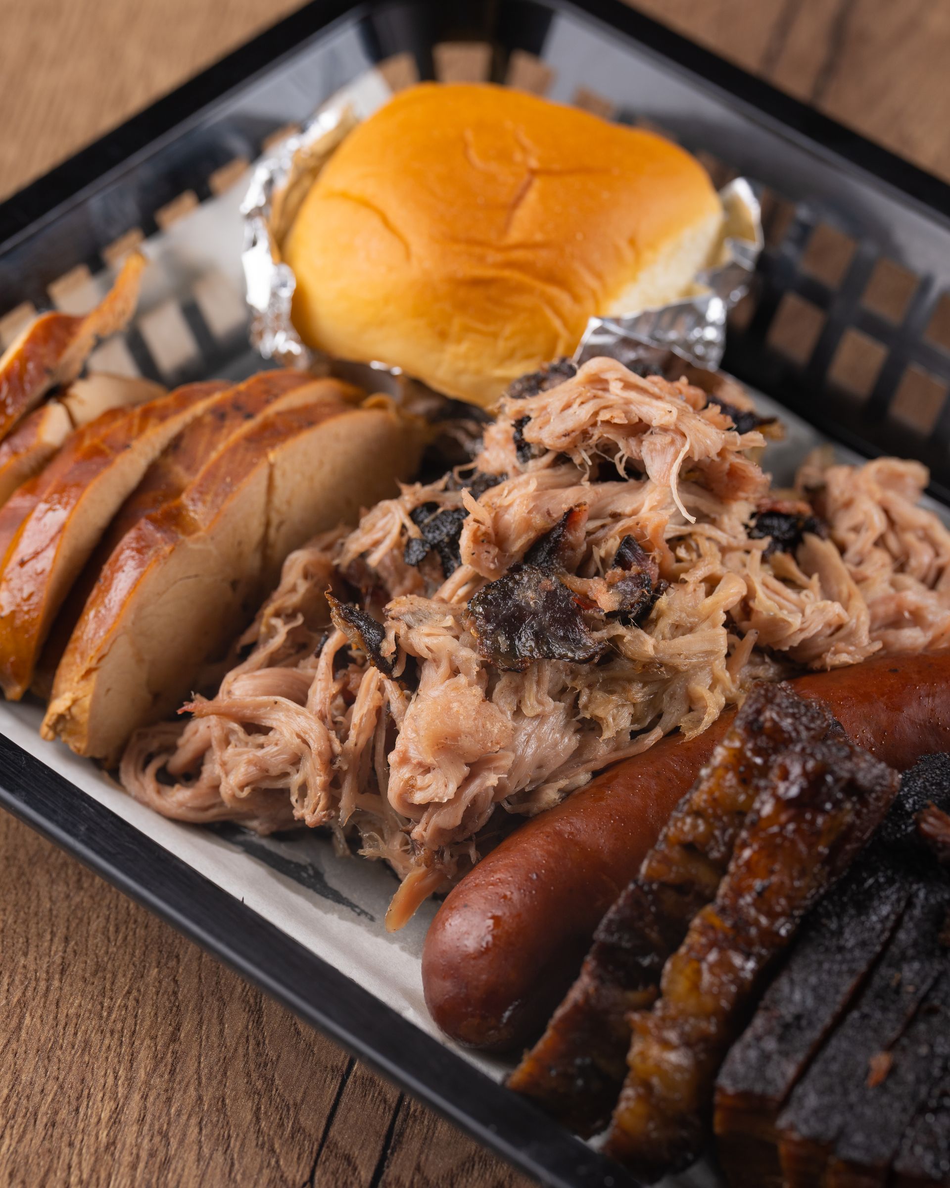 BBQ platter: sliced chicken, pulled pork, sausage, ribs, and a roll, in a black basket.