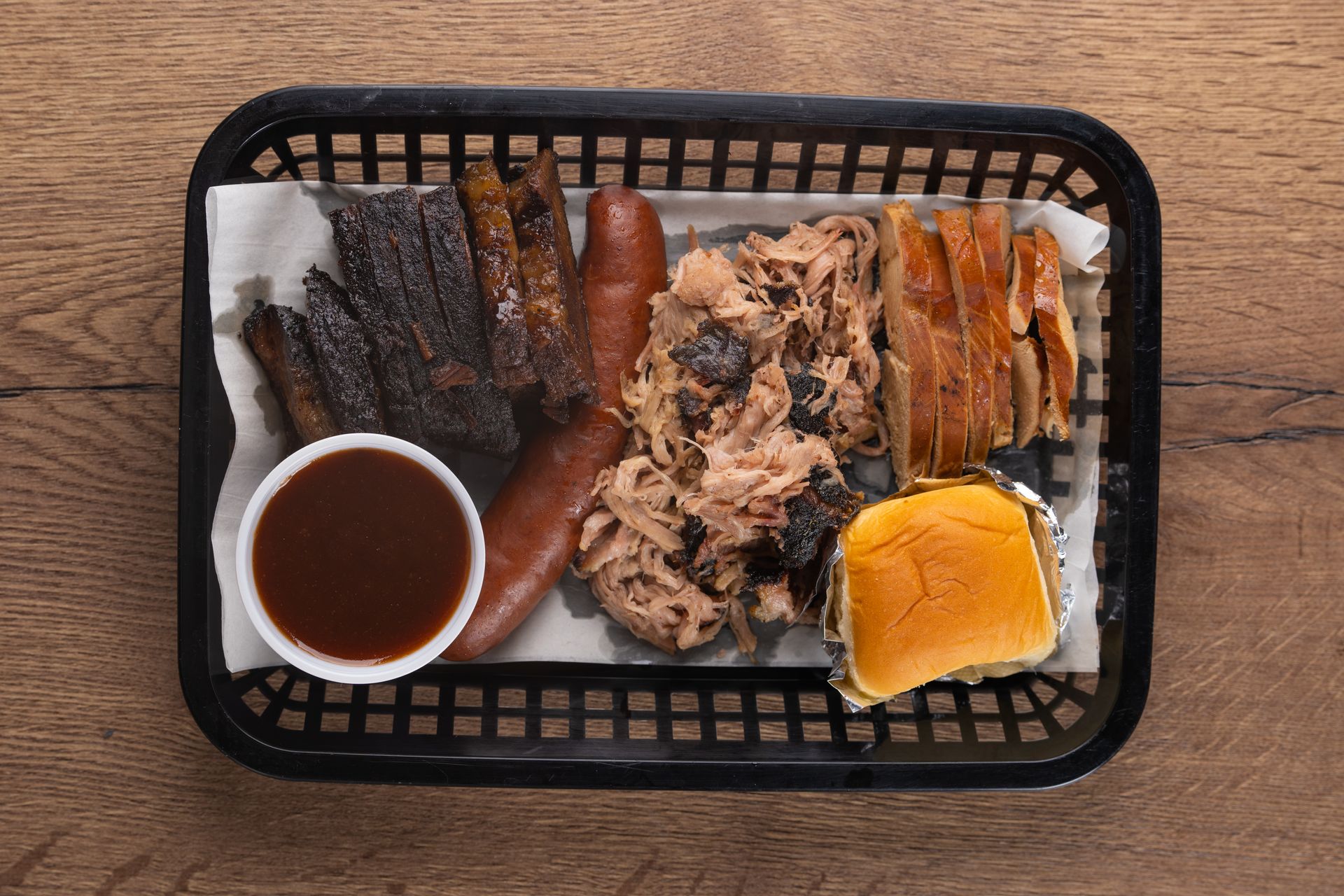 BBQ platter in a black basket with brisket, sausage, pulled pork, sauce, and a bun.