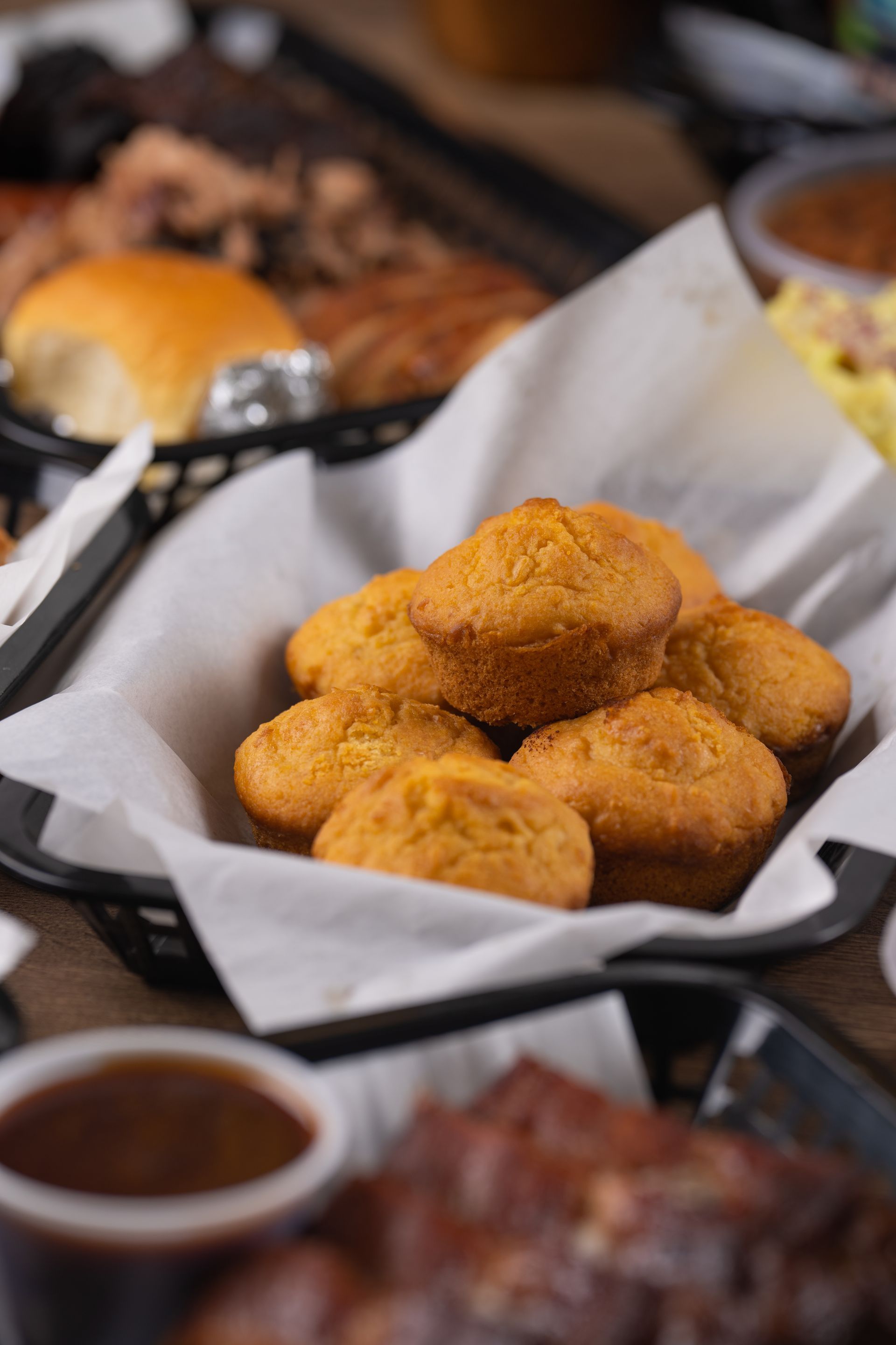 Cornbread muffins in a basket, part of a barbecue meal with other food items in the background.