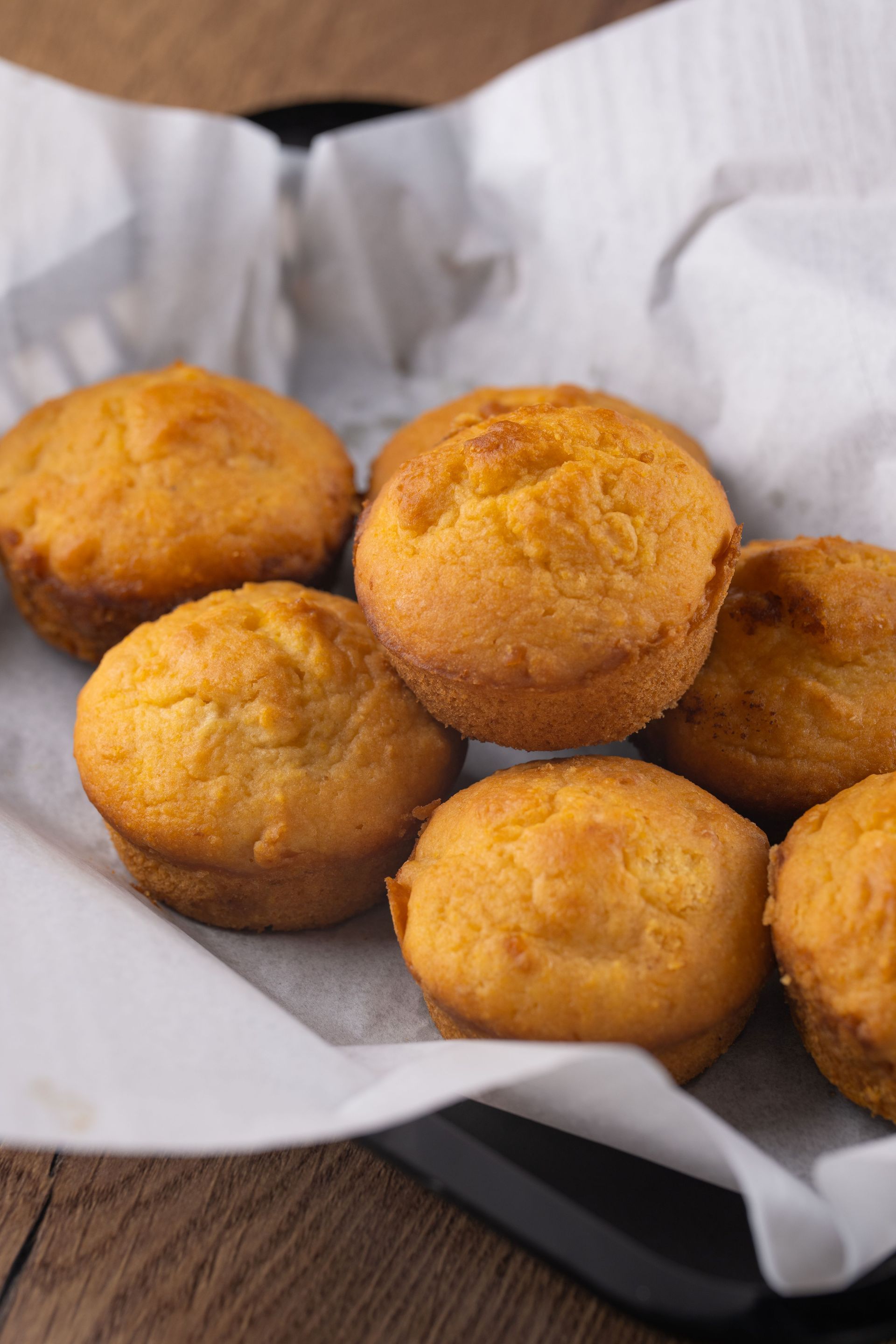 Cornbread muffins in a basket lined with white paper, on a wooden surface.