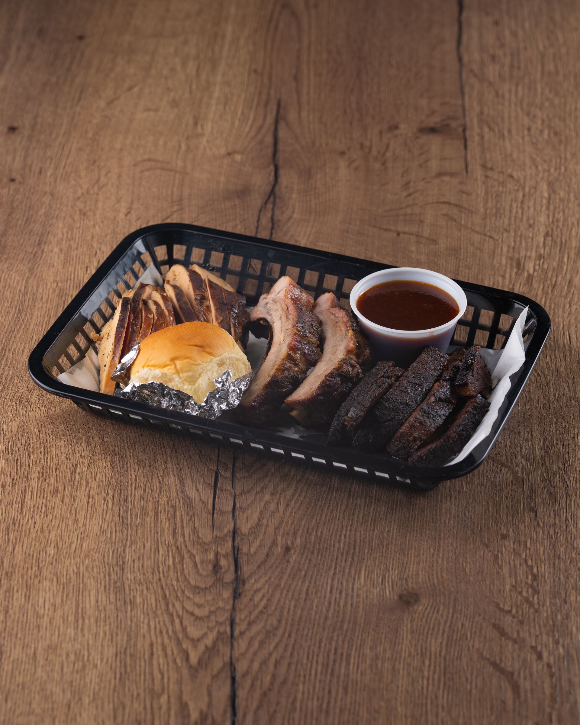 BBQ platter with sliced brisket, ribs, sauce, and a bun in a black basket on wood.