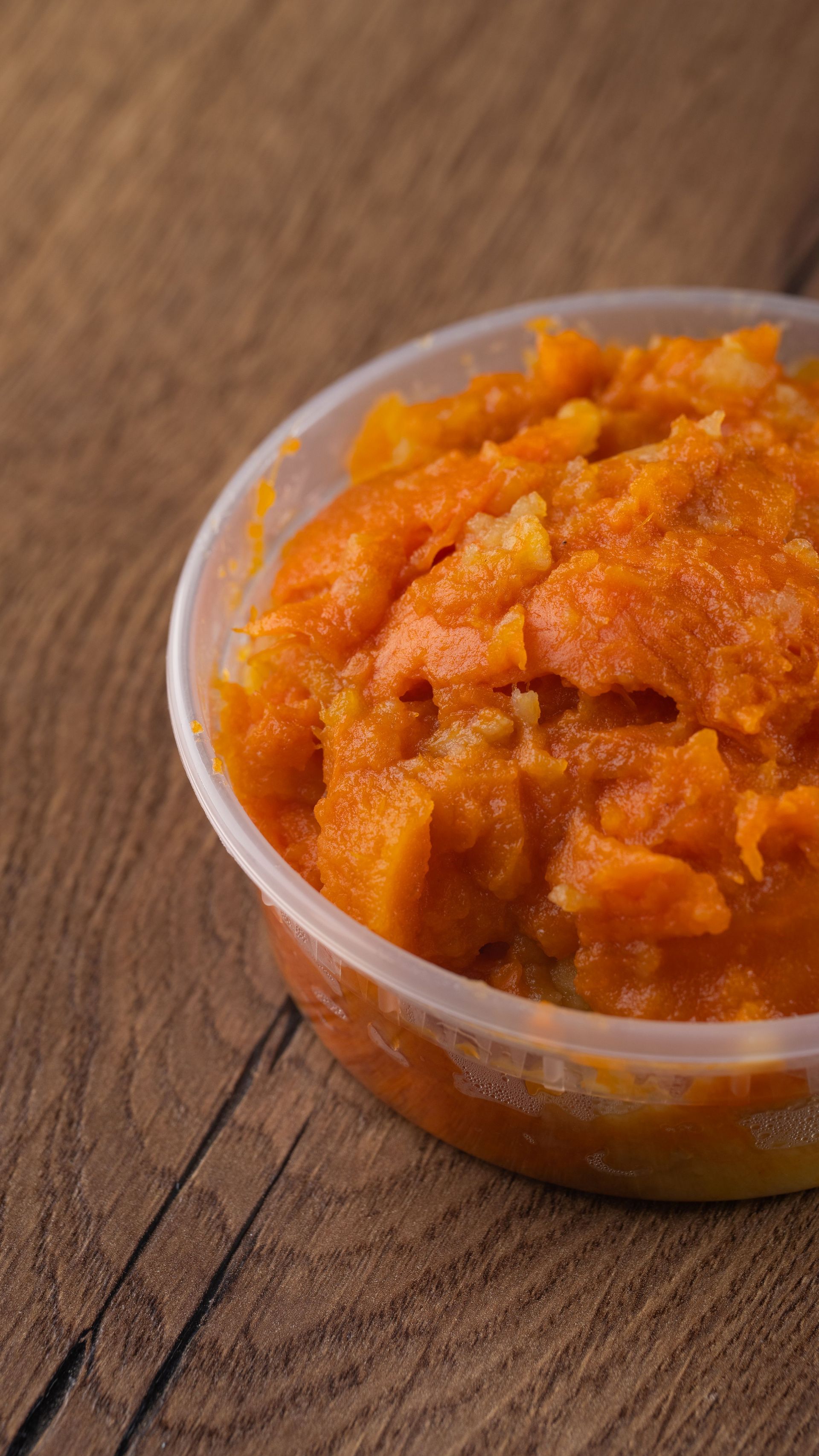 Orange-colored food, possibly mashed sweet potatoes or squash, in a clear plastic bowl on a wood table.
