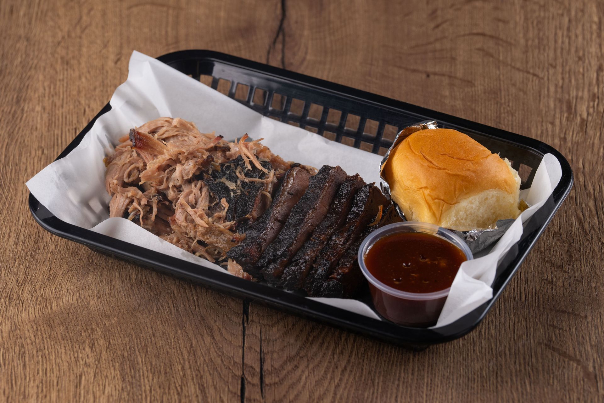 BBQ platter: Pulled pork, brisket slices, bun, and sauce in a black basket on a wood table.