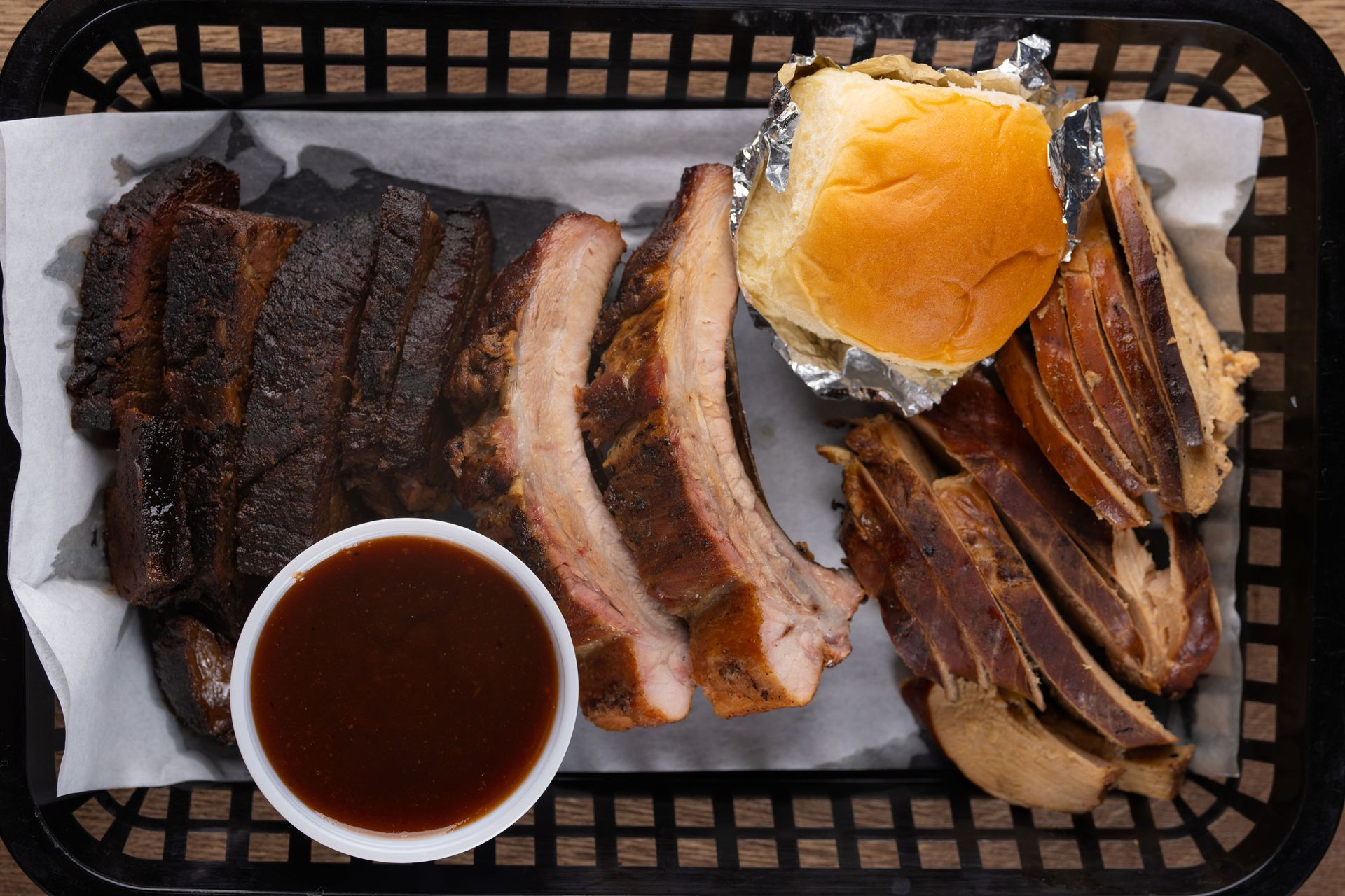 BBQ platter: ribs, sliced brisket, roll, and sauce in a black basket lined with paper.