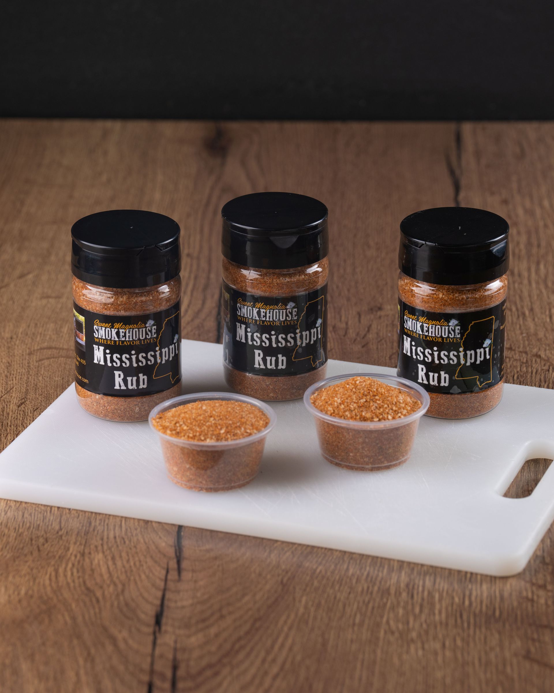 Three spice rub containers and two sample cups on a white cutting board, set on a wooden surface.