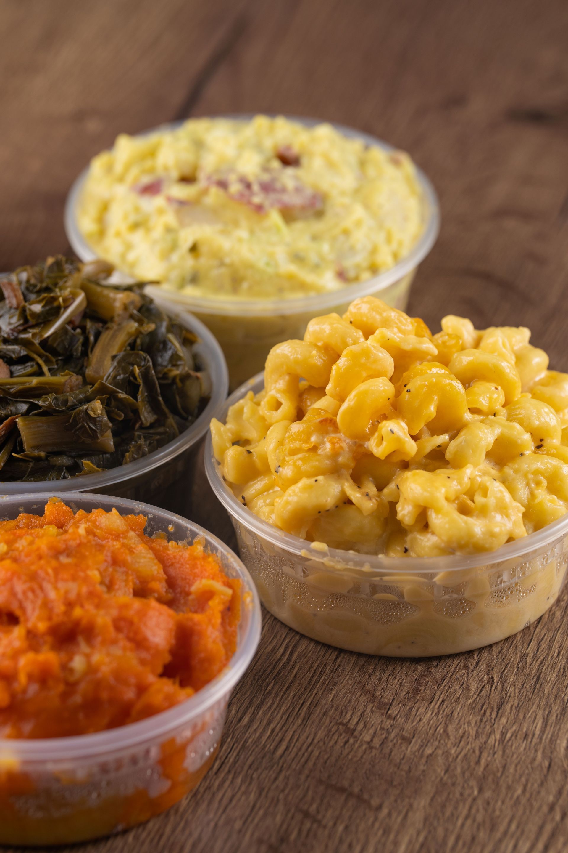 Four clear plastic containers filled with sides: mashed sweet potatoes, collard greens, mac and cheese, and potato salad.