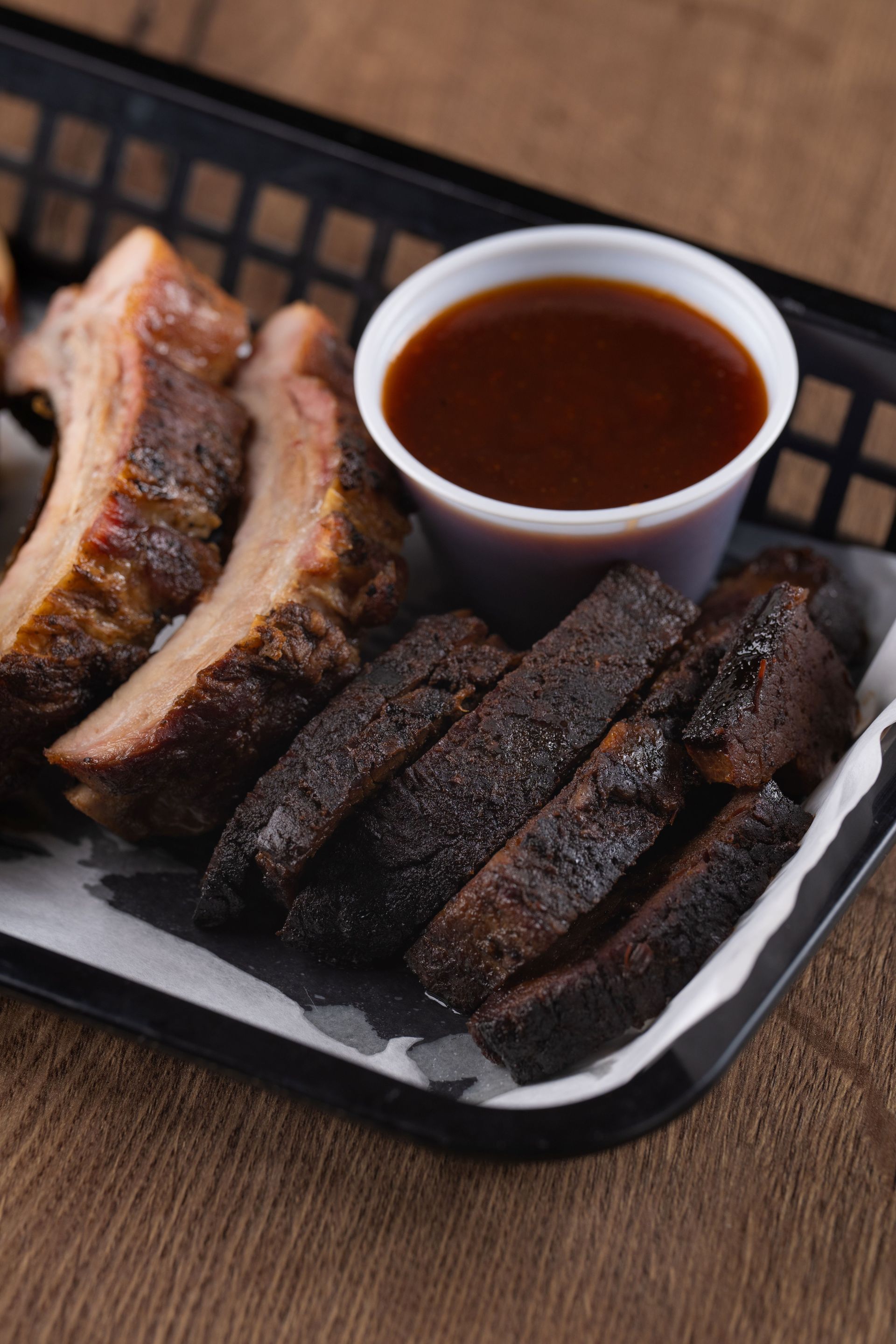 BBQ ribs, brisket, and sauce in a black basket on a wooden table.