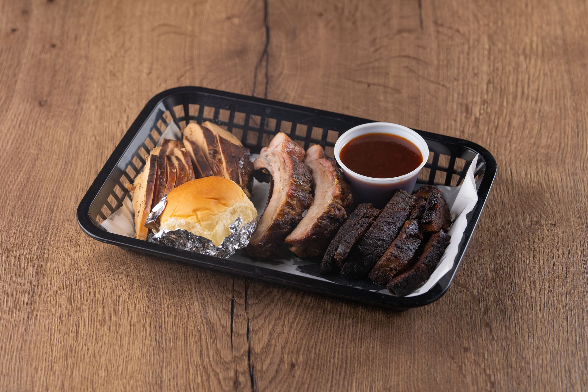 BBQ ribs, sausage, and bread in a black basket with sauce and a roll on a wooden table.