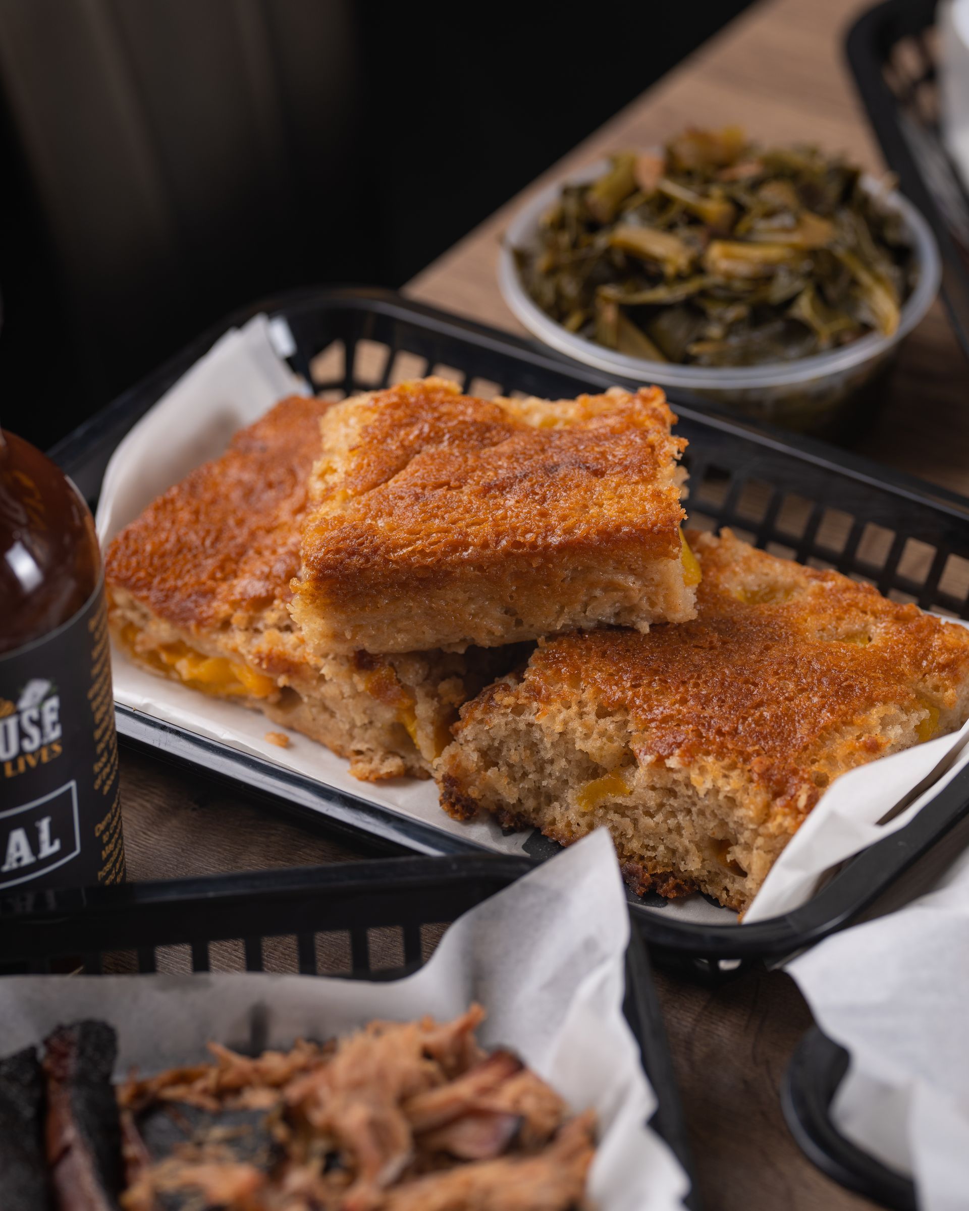 Cornbread slices with BBQ sauce and collard greens in a restaurant setting.