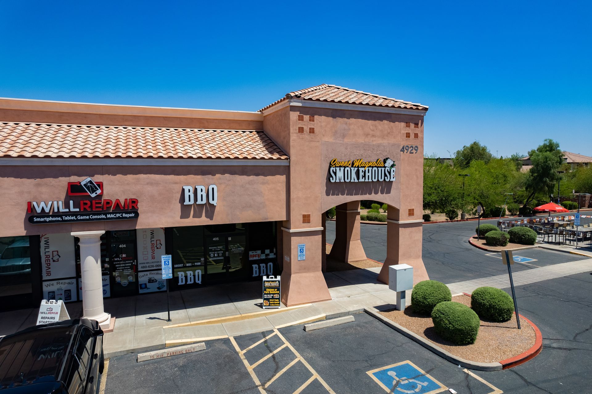 Exterior of a strip mall with a Will Repair shop and a BBQ restaurant under a bright blue sky.