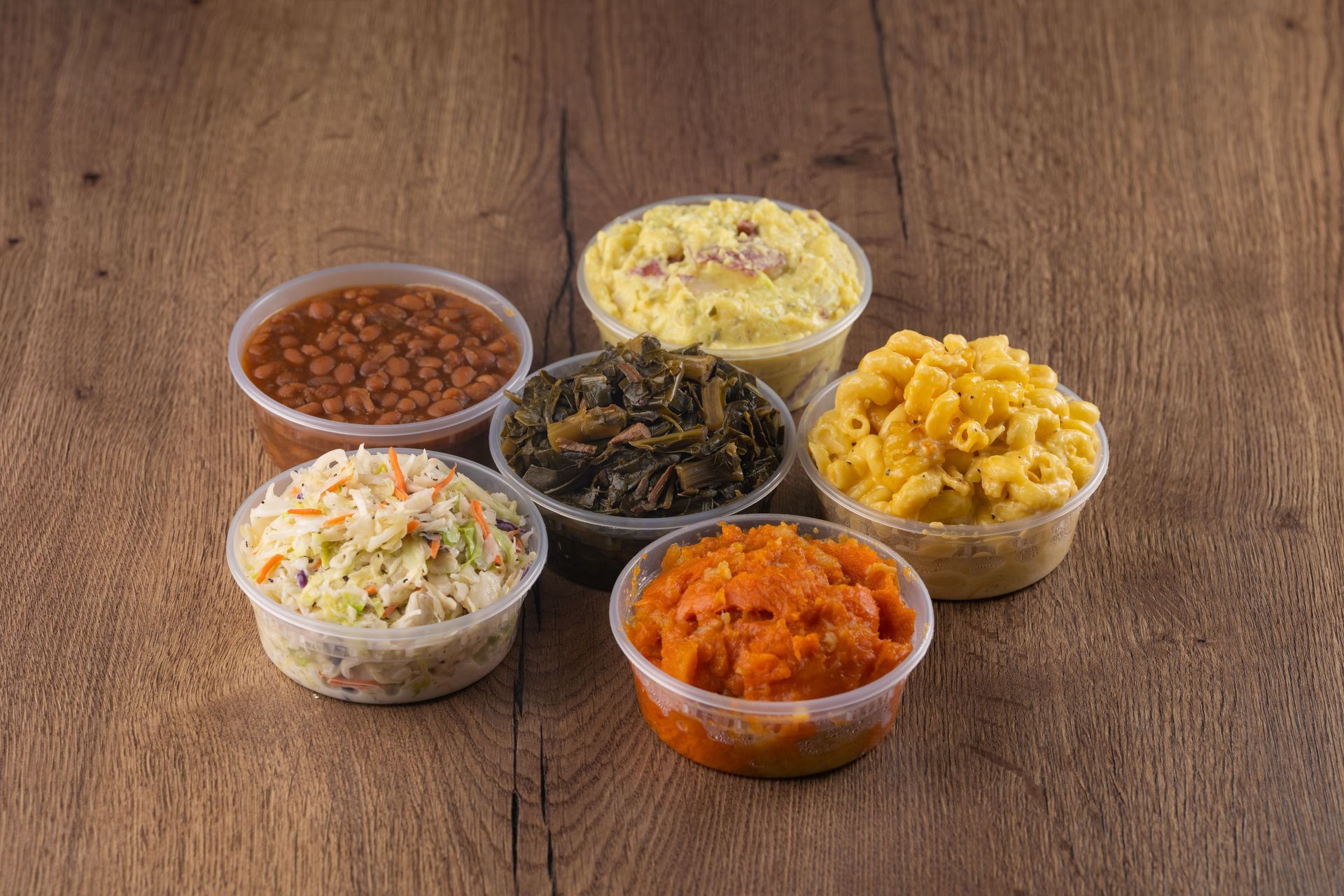 Bowls of side dishes: baked beans, potato salad, collard greens, mac and cheese, sweet potatoes, and coleslaw.