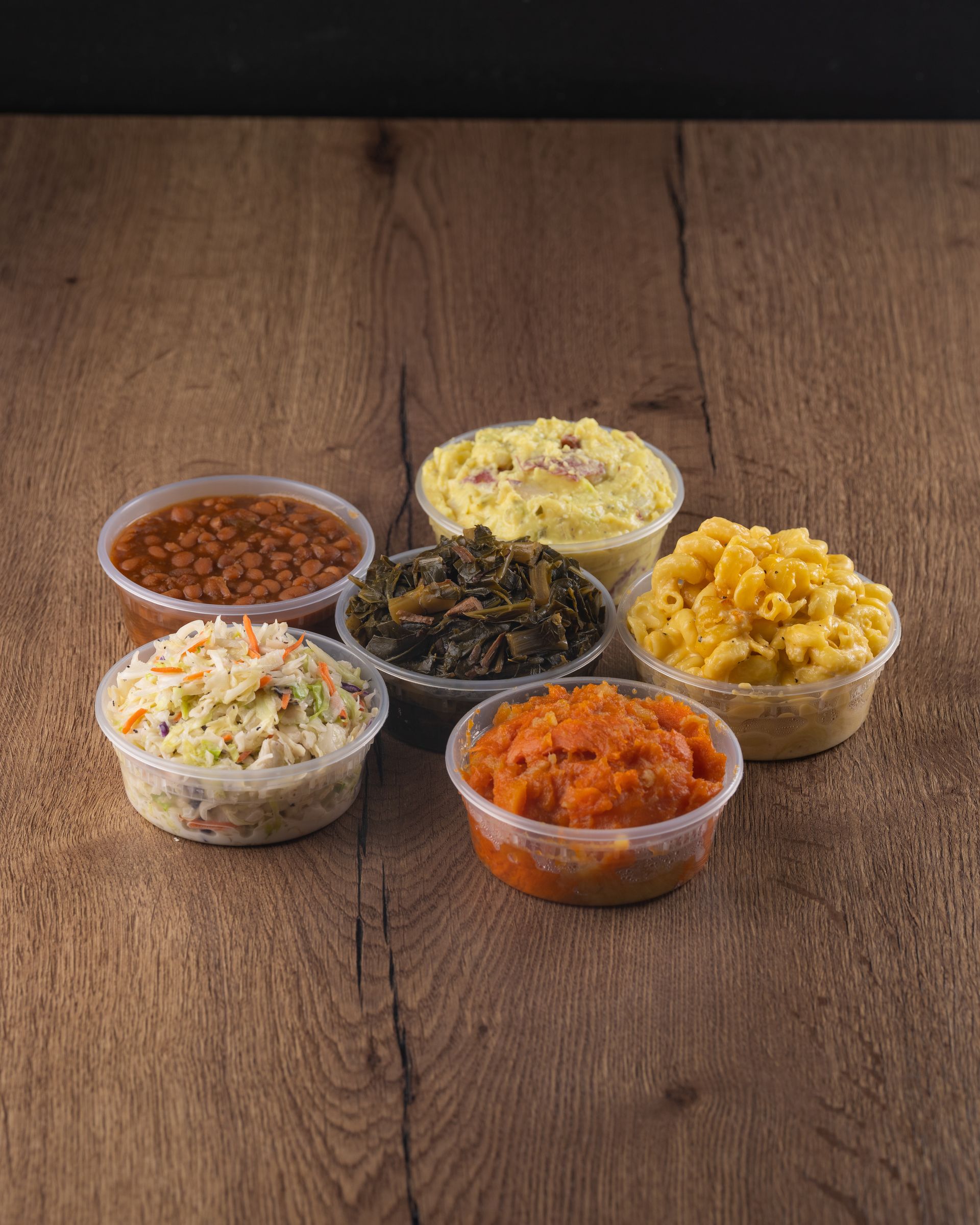Six plastic containers of sides on a wood table: beans, coleslaw, greens, potato salad, mac and cheese, carrots.