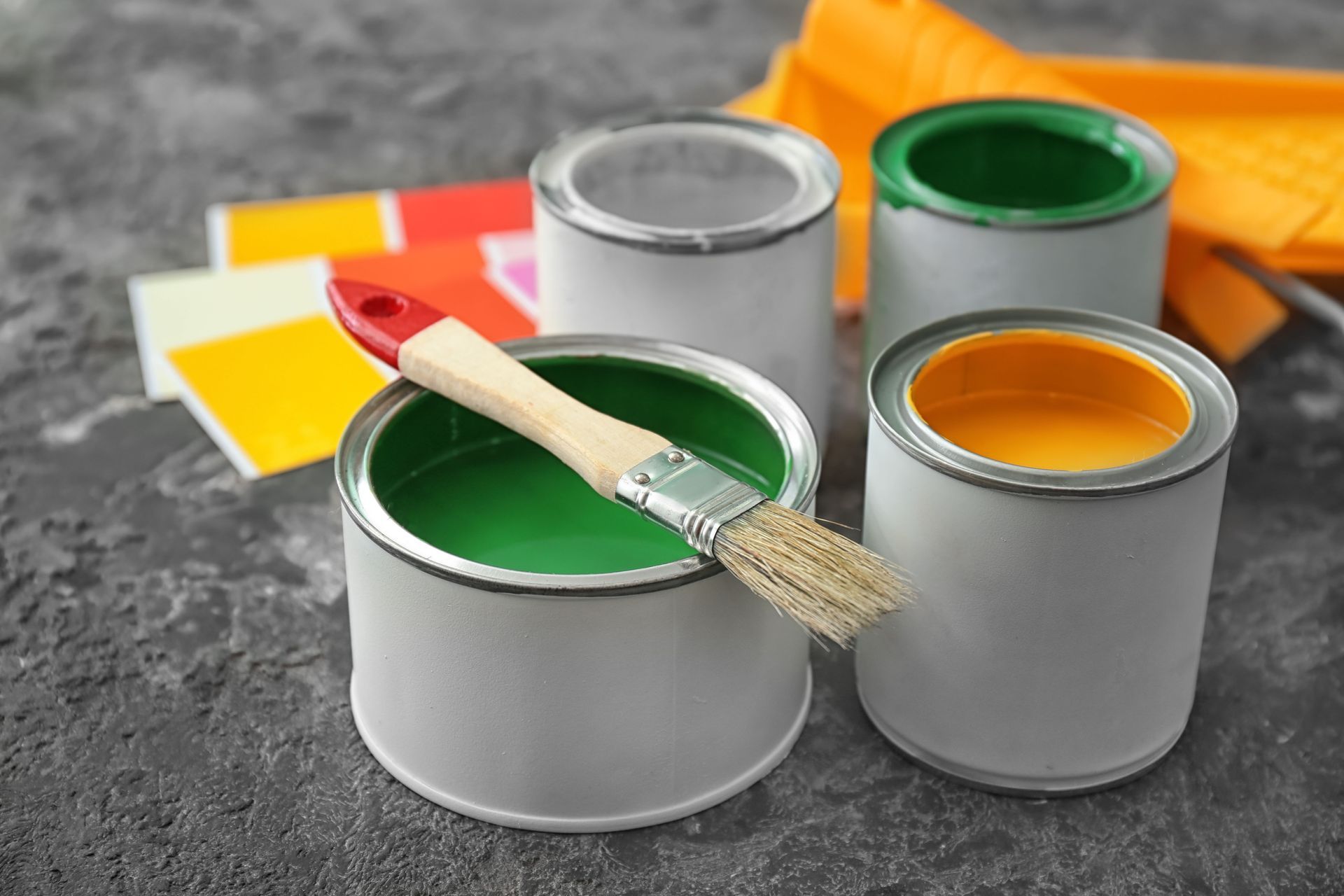 Paint cans with green, white, and yellow paint and a brush on a gray surface