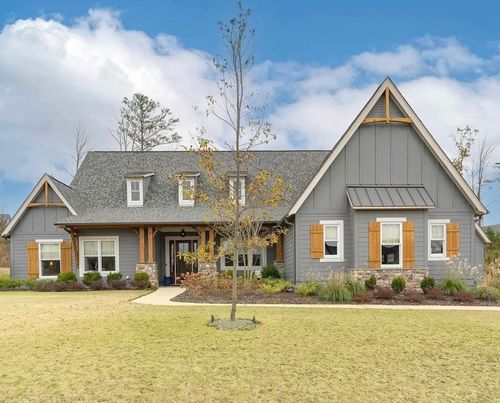 Gray suburban house with a gabled roof, front porch, and a young tree in a grassy yard under a blue sky