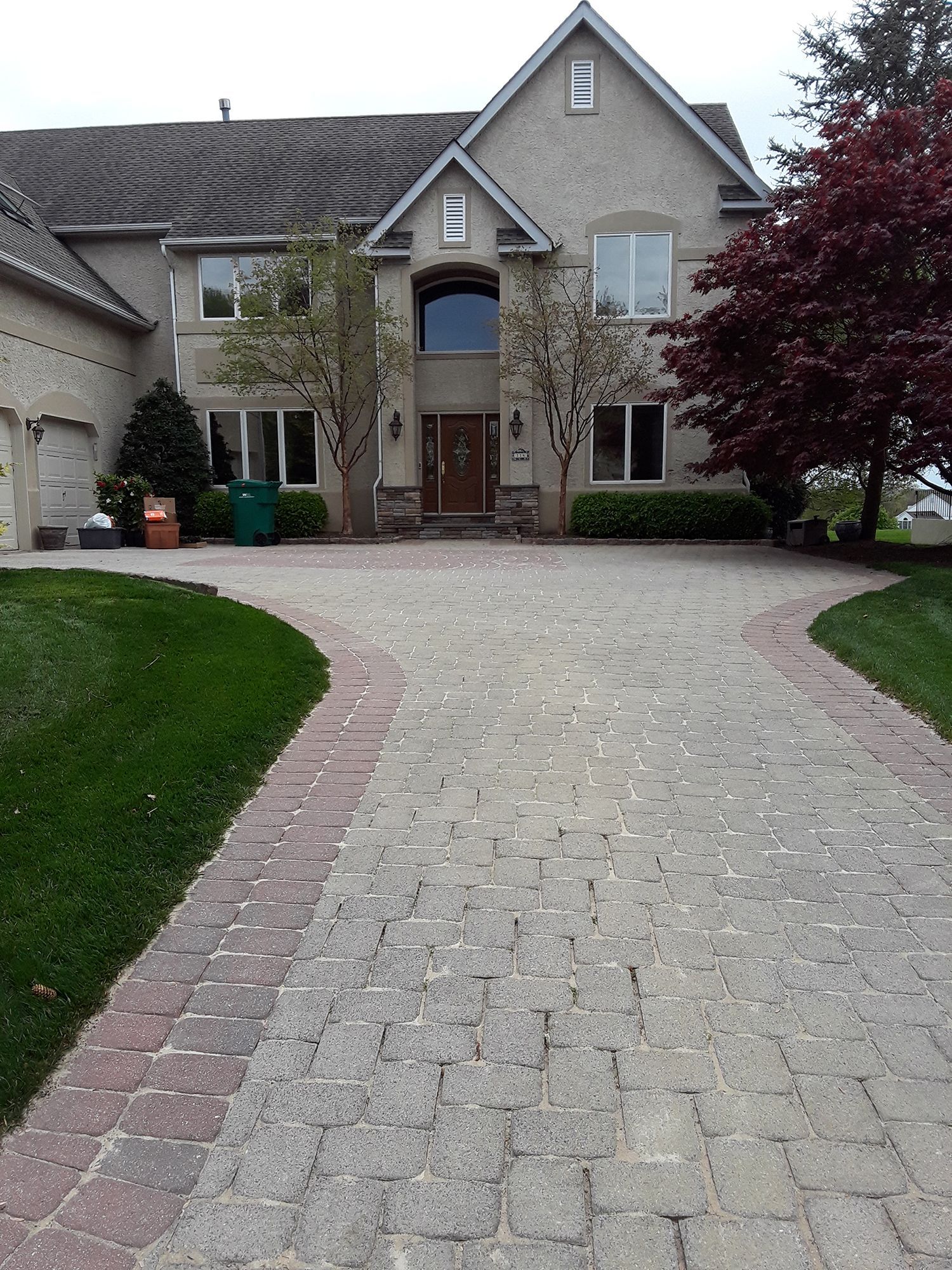 A brick driveway leading to a two-story house with a peaked roof and landscaped lawn.
