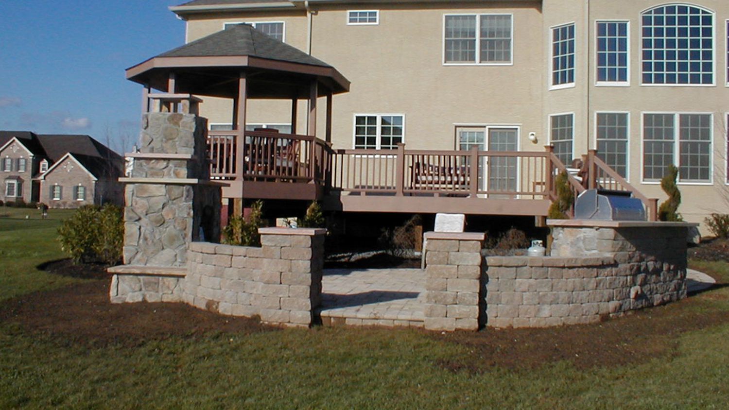 Backyard with gazebo, deck, and stone wall, next to a large house.