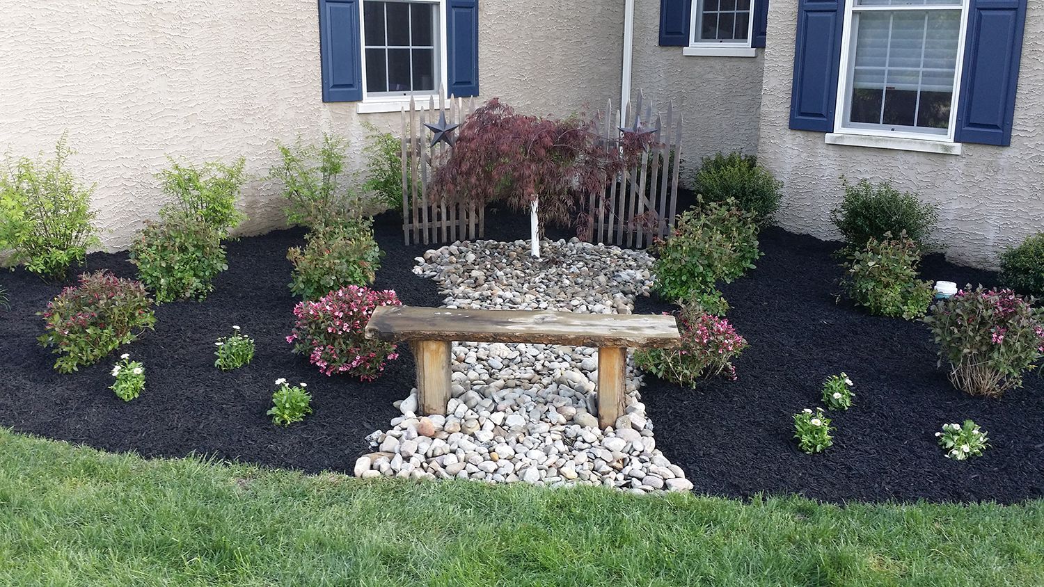 Front yard with bench and rock garden, surrounded by black mulch and bushes near a house with blue shutters.