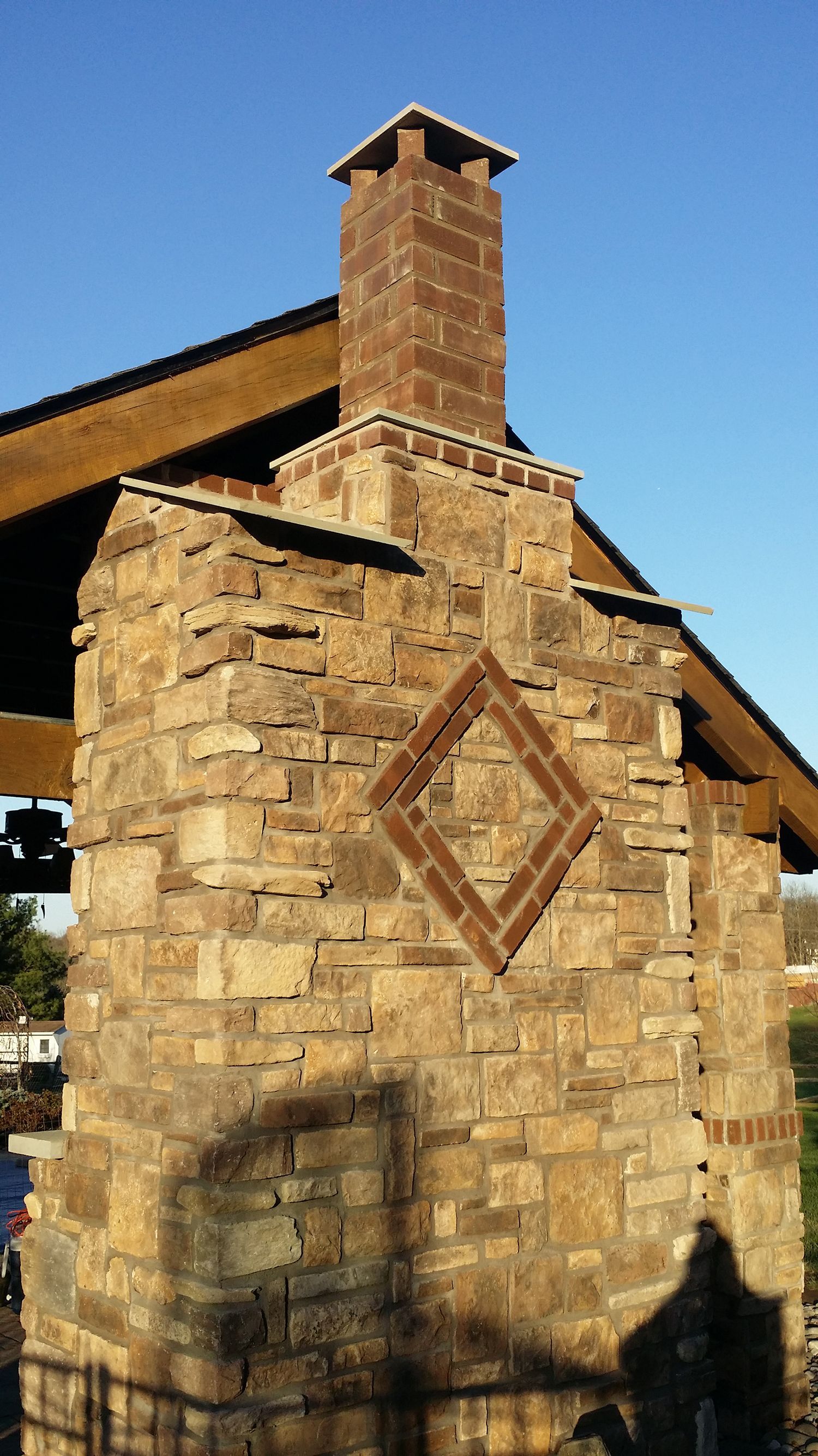 Stone chimney with brick accent and matching flue against a blue sky, beneath a wooden roof.