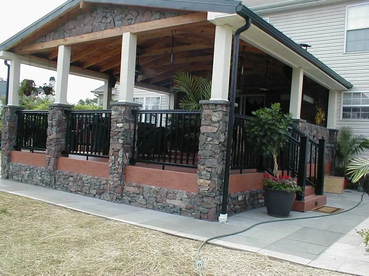 Covered porch with stone columns and black railings. House exterior with white siding and a concrete walkway.