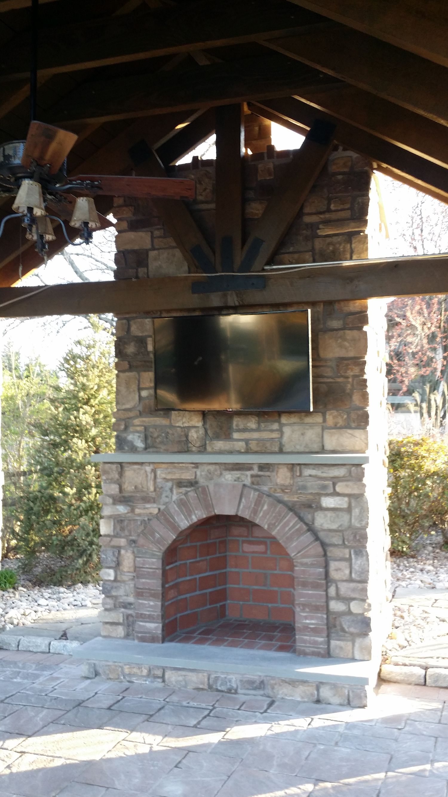 Stone fireplace with a brick firebox, a TV mounted above, under a wooden pergola.