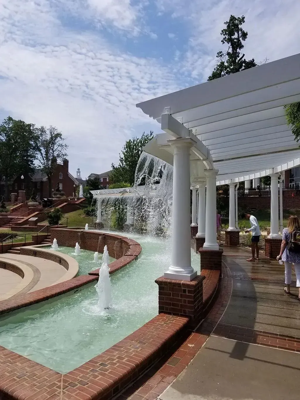 Fountain with white columns, cascading water, and brick trim under a pergola, sunny day.