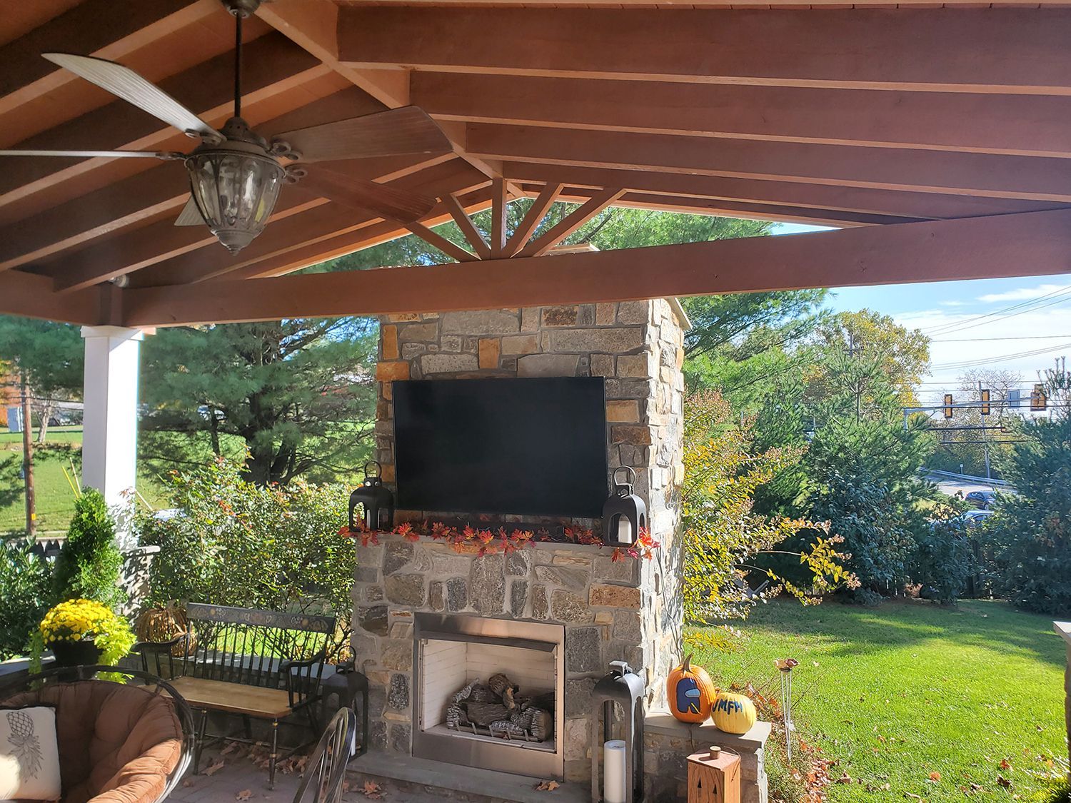 Outdoor stone fireplace with TV, ceiling fan, and surrounding greenery.