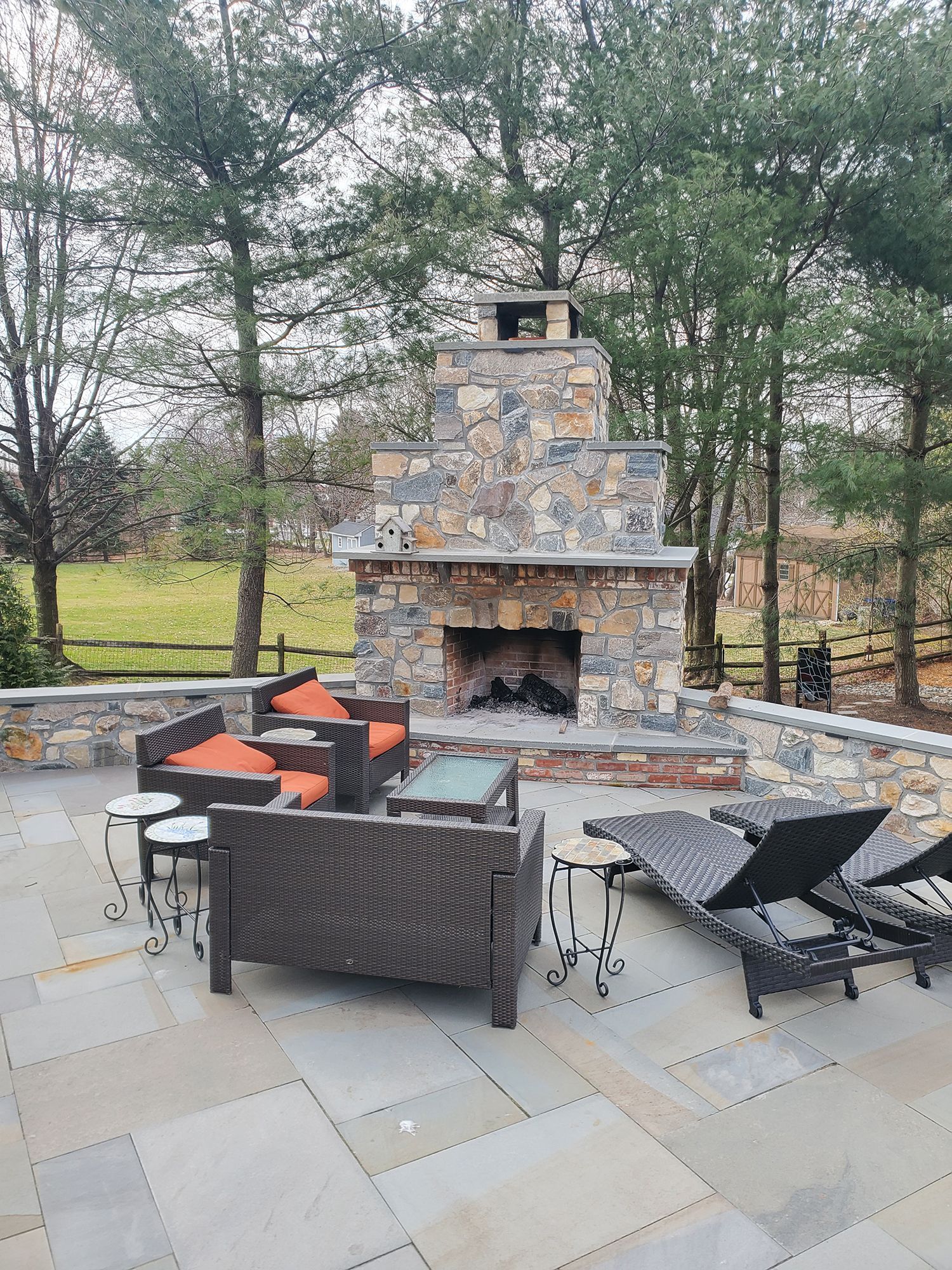 Stone outdoor fireplace on a patio with seating. Trees and green lawn in the background.