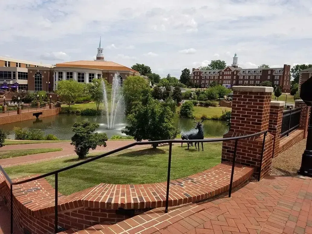 Brick steps overlook a pond with a fountain, trees, and buildings. Sunny day.