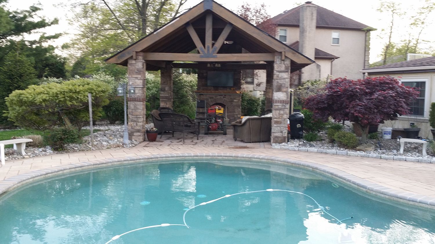 Poolside patio with stone fireplace, TV, and pavilion. Pool in the foreground, house in the background.