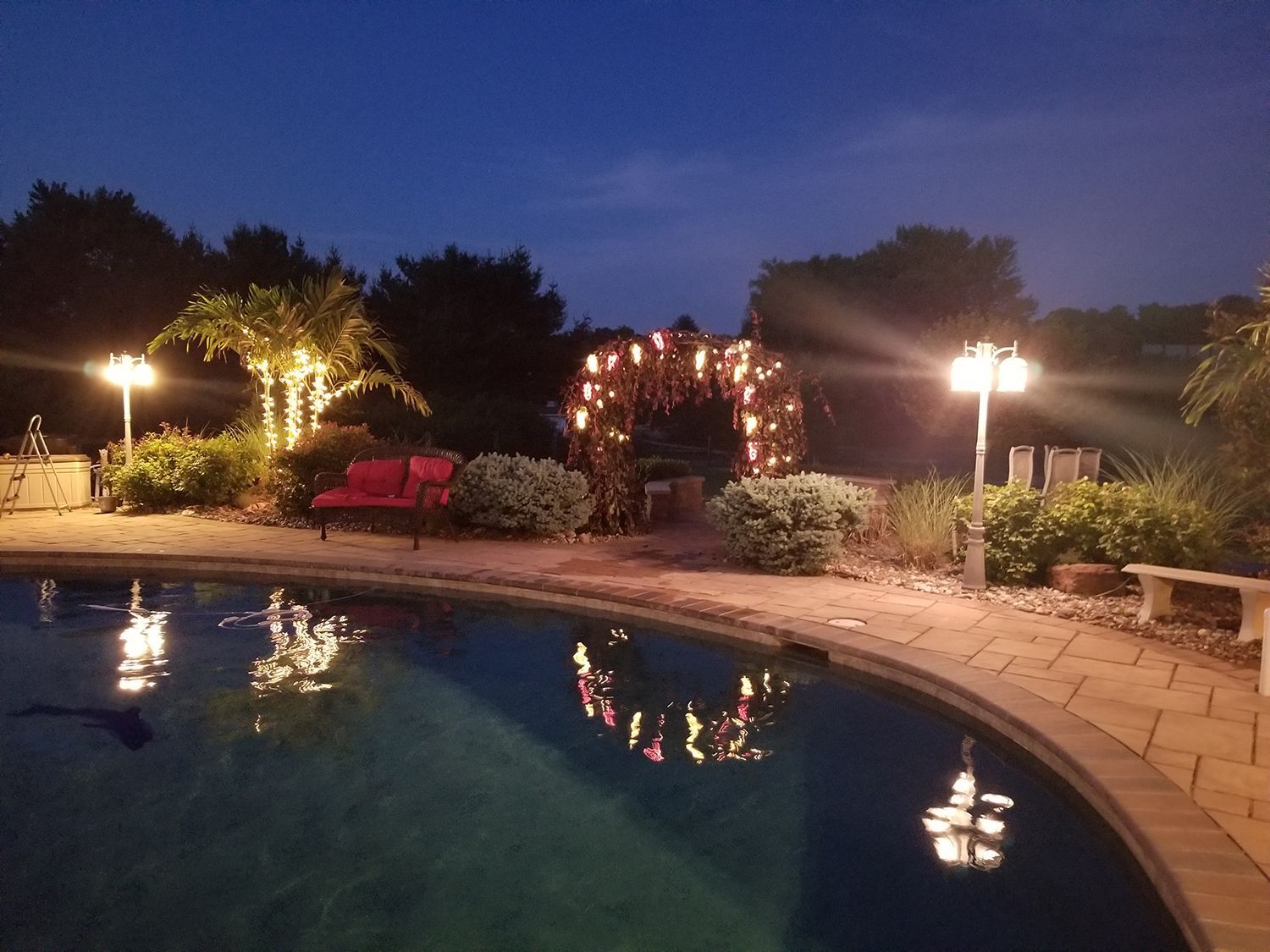 Night view of a backyard pool, illuminated by lights reflecting on the water. A red bench and decorated archway are visible.