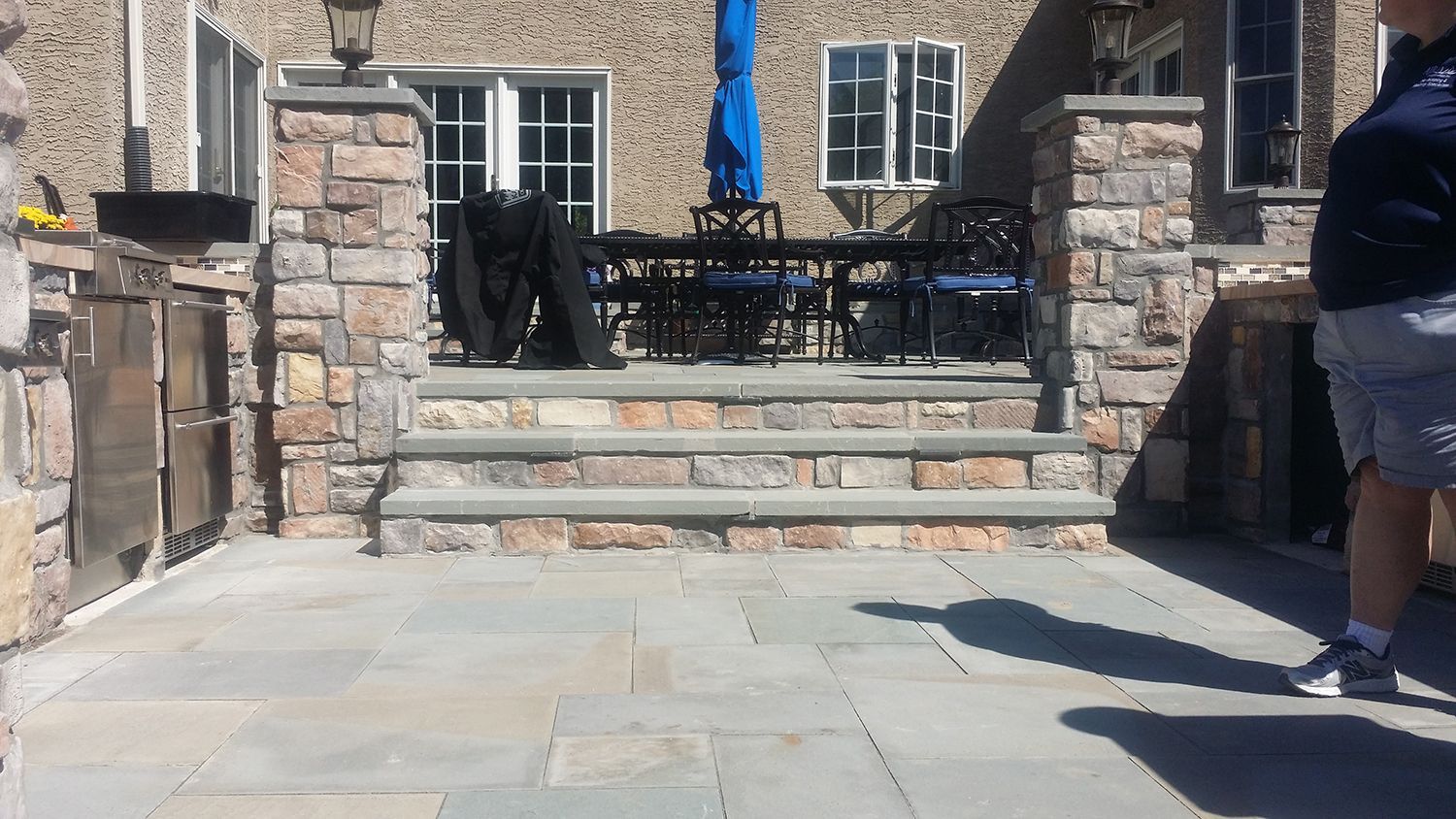 Stone patio with steps leading up to a dining area. A person stands on the right. Blue umbrella visible.