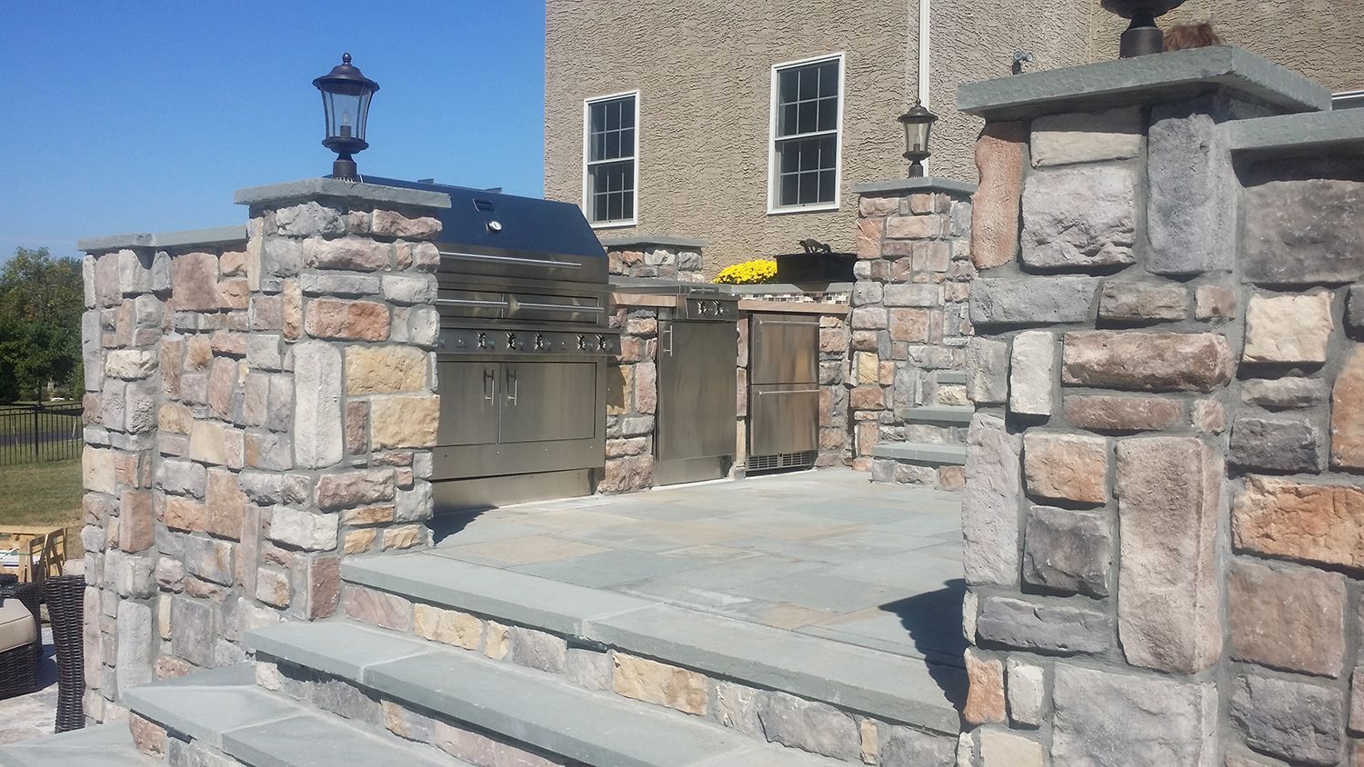 Stone outdoor kitchen with grill and cabinetry, on a stone patio with steps.